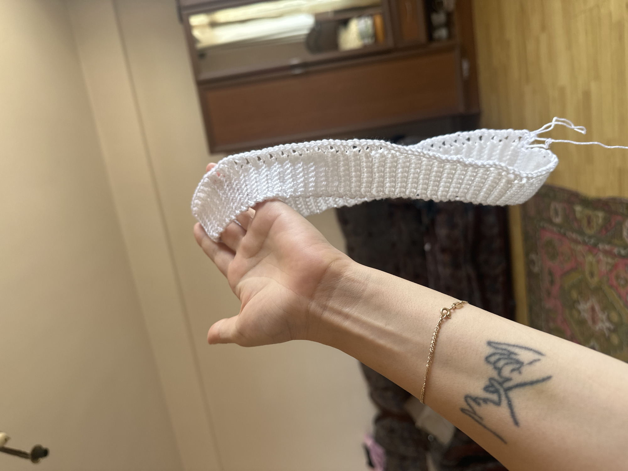 In Gyumri, Armenia, a person's hand and forearm are in the foreground, holding a piece of white crocheted fabric. The fabric has a delicate, lacy pattern along one edge and appears to be unfinished, with loose white threads hanging from the bottom. The person is wearing a thin gold chain bracelet around their wrist. A tattoo of two hands doing a pinky promise is visible on the forearm. In the background, a wooden floor, a patterned rug, and part of a closet with mirrored doors are visible. The lighting suggests an indoor environment, possibly a living room or bedroom.