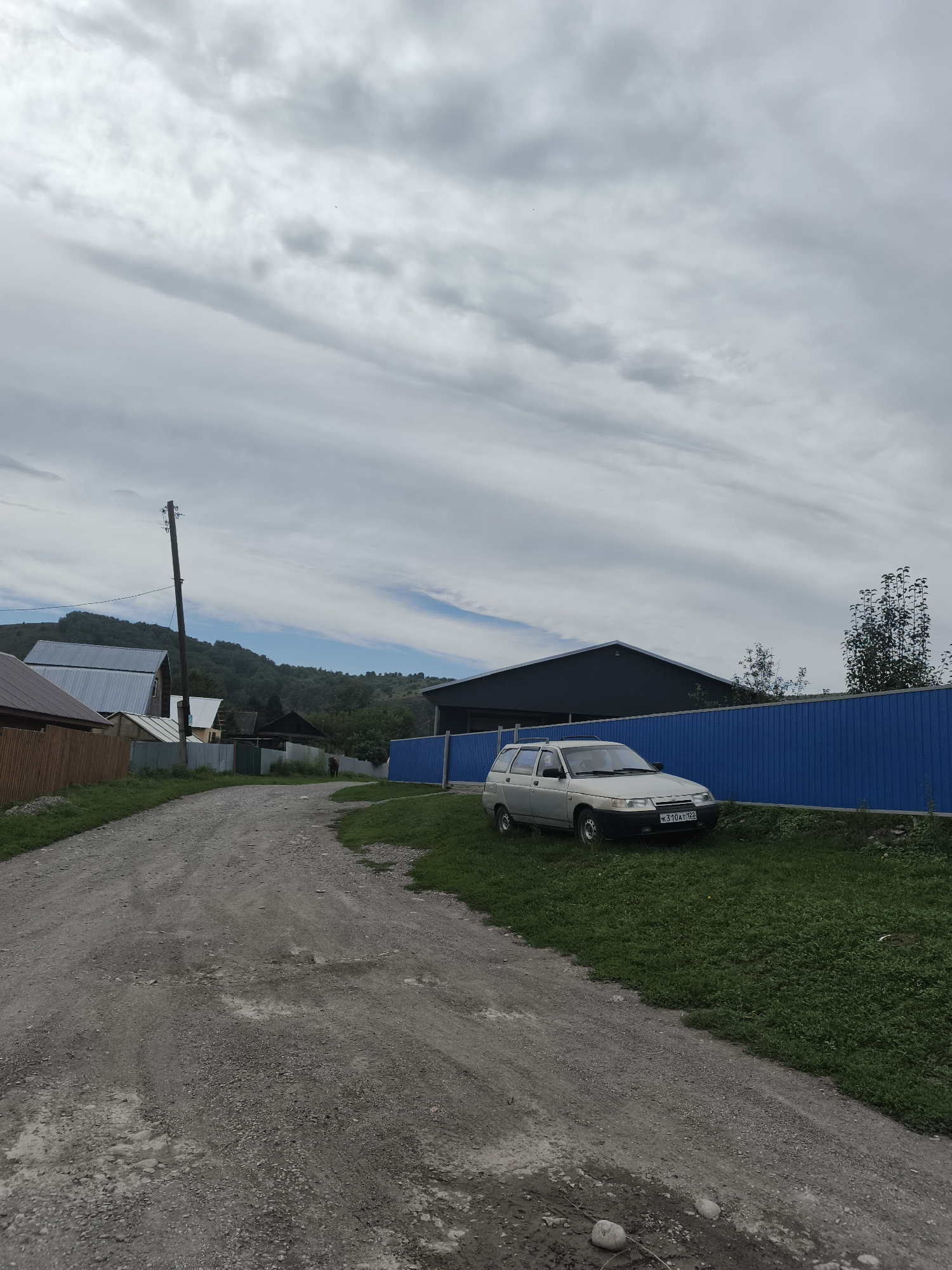 The image shows a rural road with a silver station wagon parked on the right side, next to a blue metal fence. The car is a Lada. The road is unpaved and appears to be made of gravel. There are houses and trees visible in the background, as well as a gently sloping hill covered in vegetation. The sky is cloudy with some patches of blue.