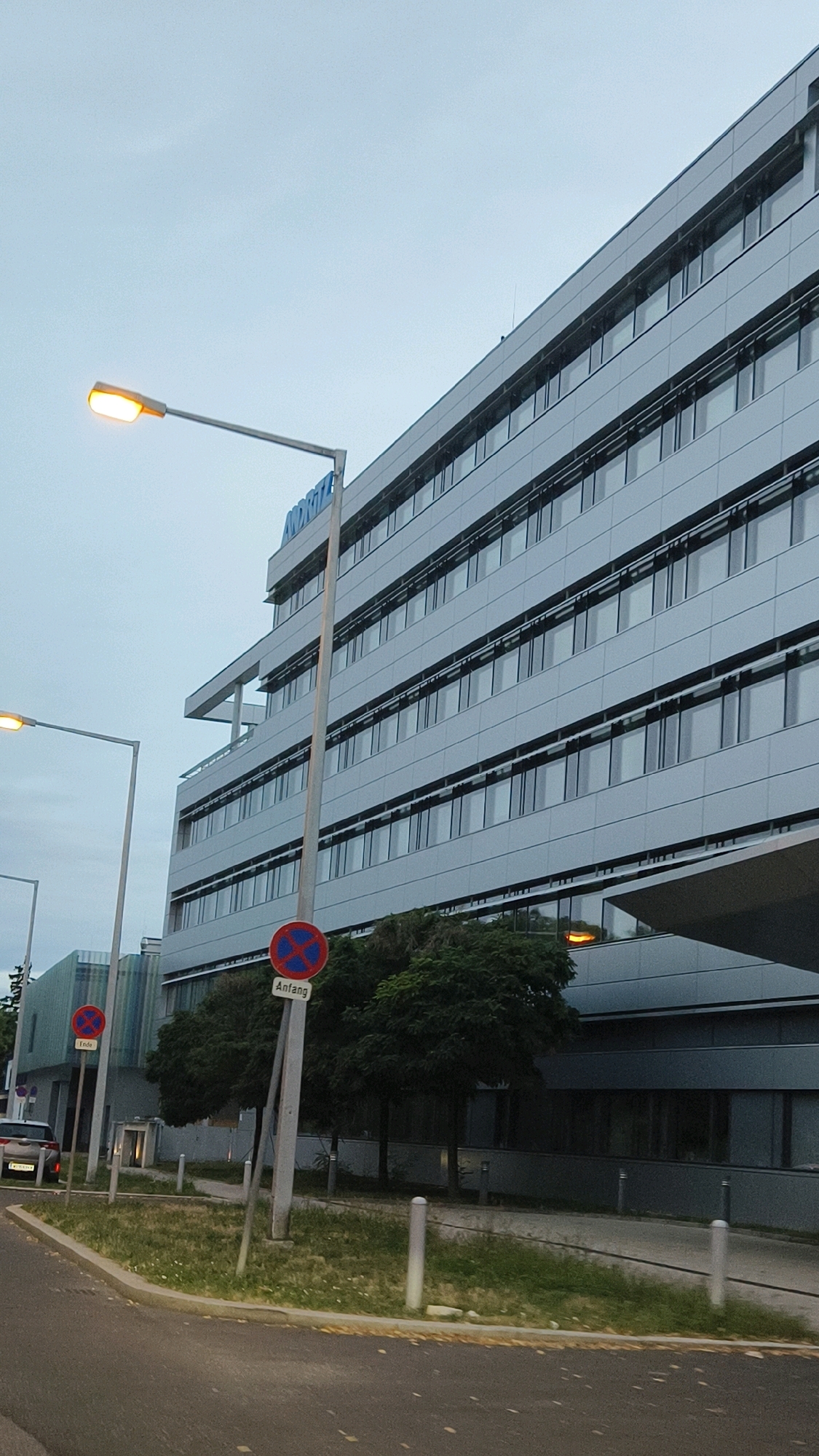 The image depicts a modern, multi-story office building with a gray, paneled facade and numerous windows. The building is located in Vienna, Austria, as indicated by the signage on a street sign that reads "Anfang" (German for "beginning"). A street lamp with a yellow light is visible in the foreground, casting a warm glow against the overcast sky. Two red and blue circular traffic signs are mounted on a pole near the building. One sign has an "X" within a red circle, indicating no stopping, and the word "Anfang" below it. Another similar sign, smaller and partially visible, has the word "Ende" (German for "end") below it. The building's architecture is sleek and contemporary, with horizontal lines formed by the window frames and facade panels. Trees with green foliage are planted in front of the building, and a grassy area with scattered white flowers is present along the pathway. In the background, a white car is parked on the side of the road. The overall scene suggests an urban environment with commercial or office spaces.