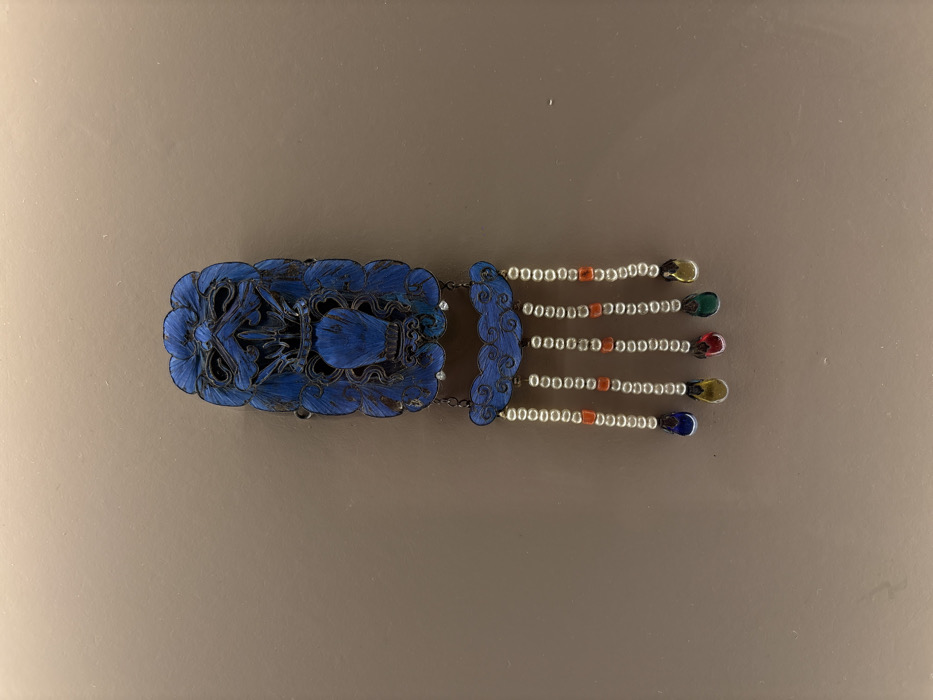 An ornate, elongated decorative object, approximately 30-35cm in length, is centrally displayed against a plain, matte light brown background. The object is primarily a deep, rich blue with black outlining and intricate surface texture, possibly indicative of enamel or feathered ornamentation. The upper section, comprising roughly two-thirds of the object, features dense, stylized motifs resembling floral patterns or a grotesque face, with a prominent central drop-shaped element. Below this, a smaller, horizontally oriented, cloud-shaped segment in matching blue is suspended by short chains. From this cloud-shaped segment, five individual strands descend vertically. Each strand consists of numerous small, spherical white beads, interspersed with fewer, smaller, spherical reddish-orange beads. The strands terminate with larger, teardrop-shaped ornaments, which from left to right are dark blue, yellow, red, green, and yellow. The illumination suggests an indoor display setting. No individuals, actions, or interactions are visible within the frame. The specific location within Mexico City, Mexico, cannot be determined from the provided image.