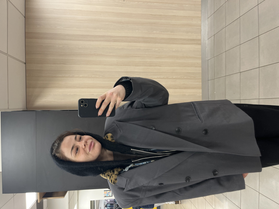 The image depicts a woman taking a mirror selfie inside a commercial establishment in Urvillers, France. The subject, a female with light brown hair and a subtle smile, occupies the central frame, looking directly into the smartphone held in her left hand. She is wearing an oversized dark grey double-breasted blazer with black buttons, a dark knitted hood with a leopard print trim, and a zippered dark top underneath. Her fingernails are painted a light color with a subtle pattern. The indoor environment features a dark vertical panel on the left, a light wood-paneled wall on the right, a white square-tiled ceiling, and a light grey square-tiled floor. Distant reflections of what appear to be clothing racks or store fixtures are faintly visible in the background.