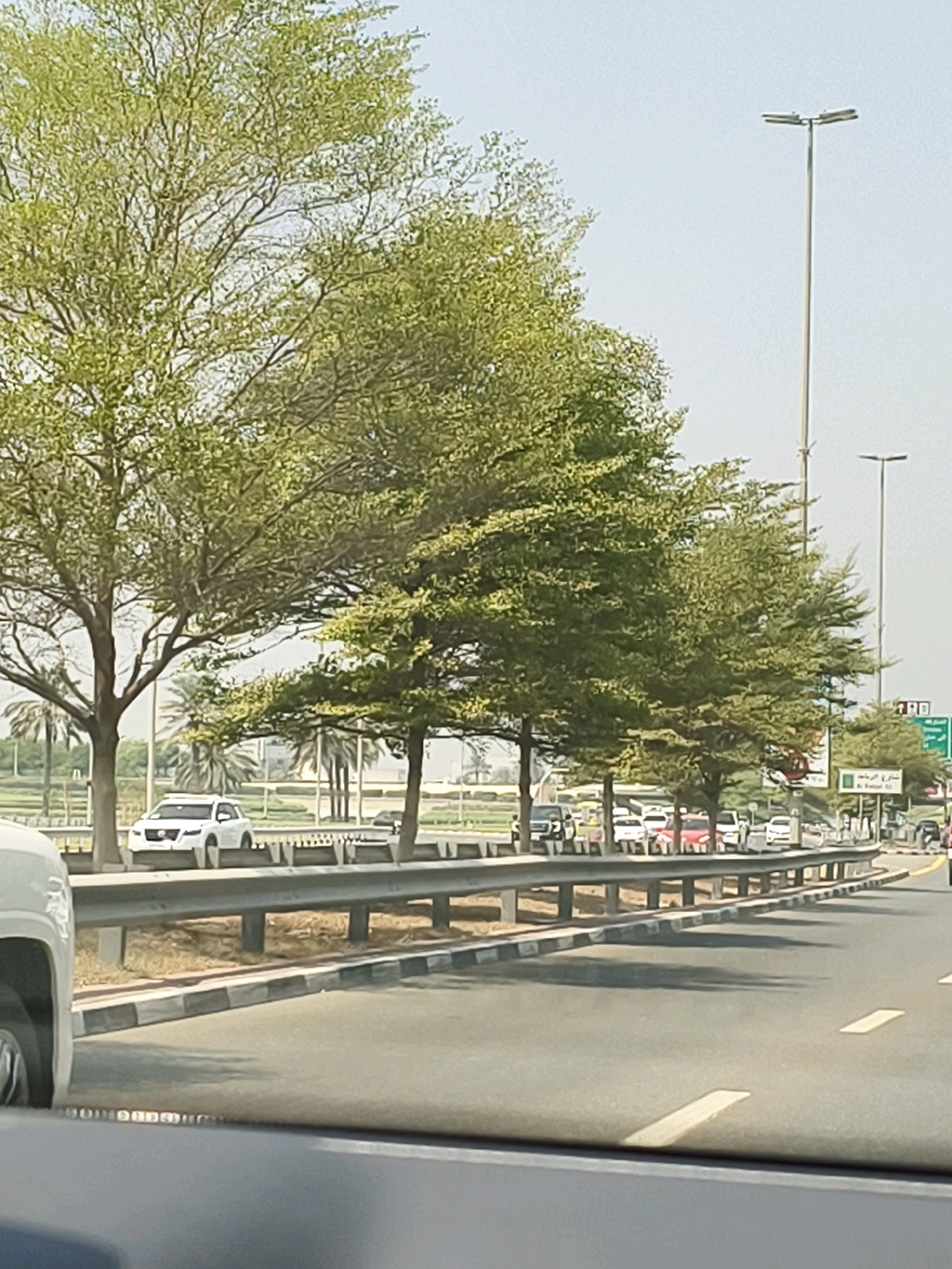 The image shows a view from a vehicle driving on a highway in Dubai, United Arab Emirates. The highway is lined with trees and palm trees. There are several cars on the highway, including a white SUV in the foreground. In the background, there are tall streetlights and road signs. One sign is in Arabic and English, indicating "Al Barsha 2". The sky is clear and the weather appears to be sunny.