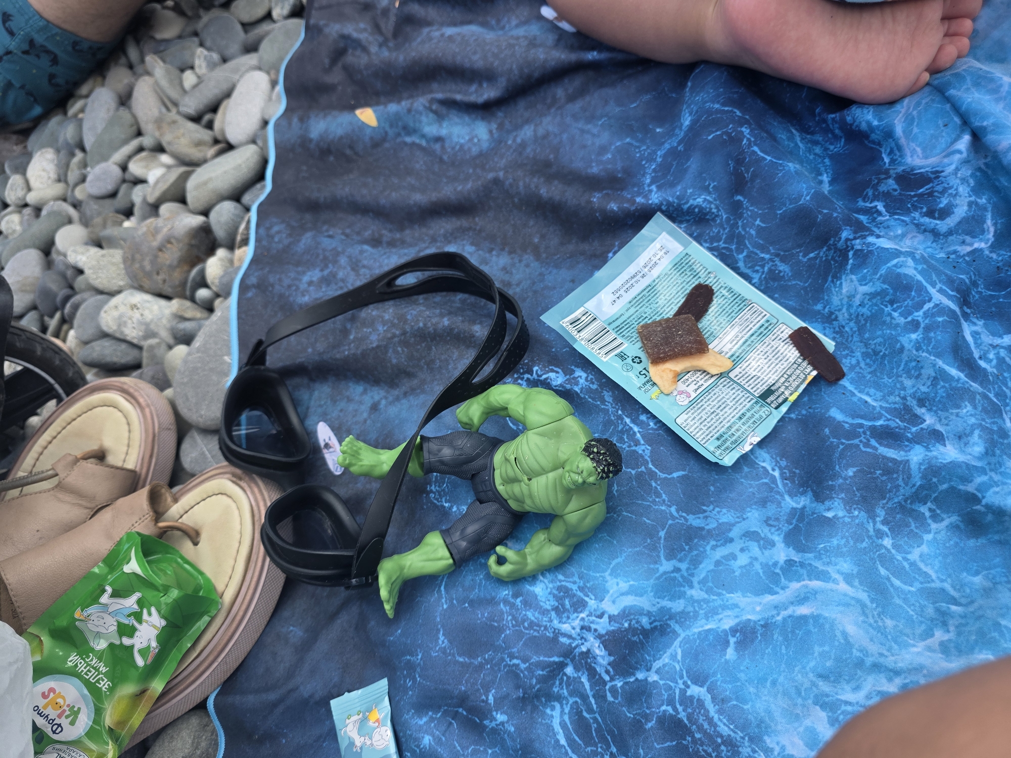 The image depicts a beach scene in Sochi, Russia. On a blue, wavy-patterned beach towel, a green Hulk action figure is lying on its back. Next to the action figure, a pair of black swimming goggles is placed. To the left of the action figure, there are two small packets, one green with cartoon animals and the other teal with text. A pair of tan flip-flops is situated behind the green packet. The background consists of smooth, grey pebbles, characteristic of a pebble beach. A person's bare foot is visible on the right side of the towel, suggesting the presence of a person. The overall setting is a relaxed outdoor environment by the sea.