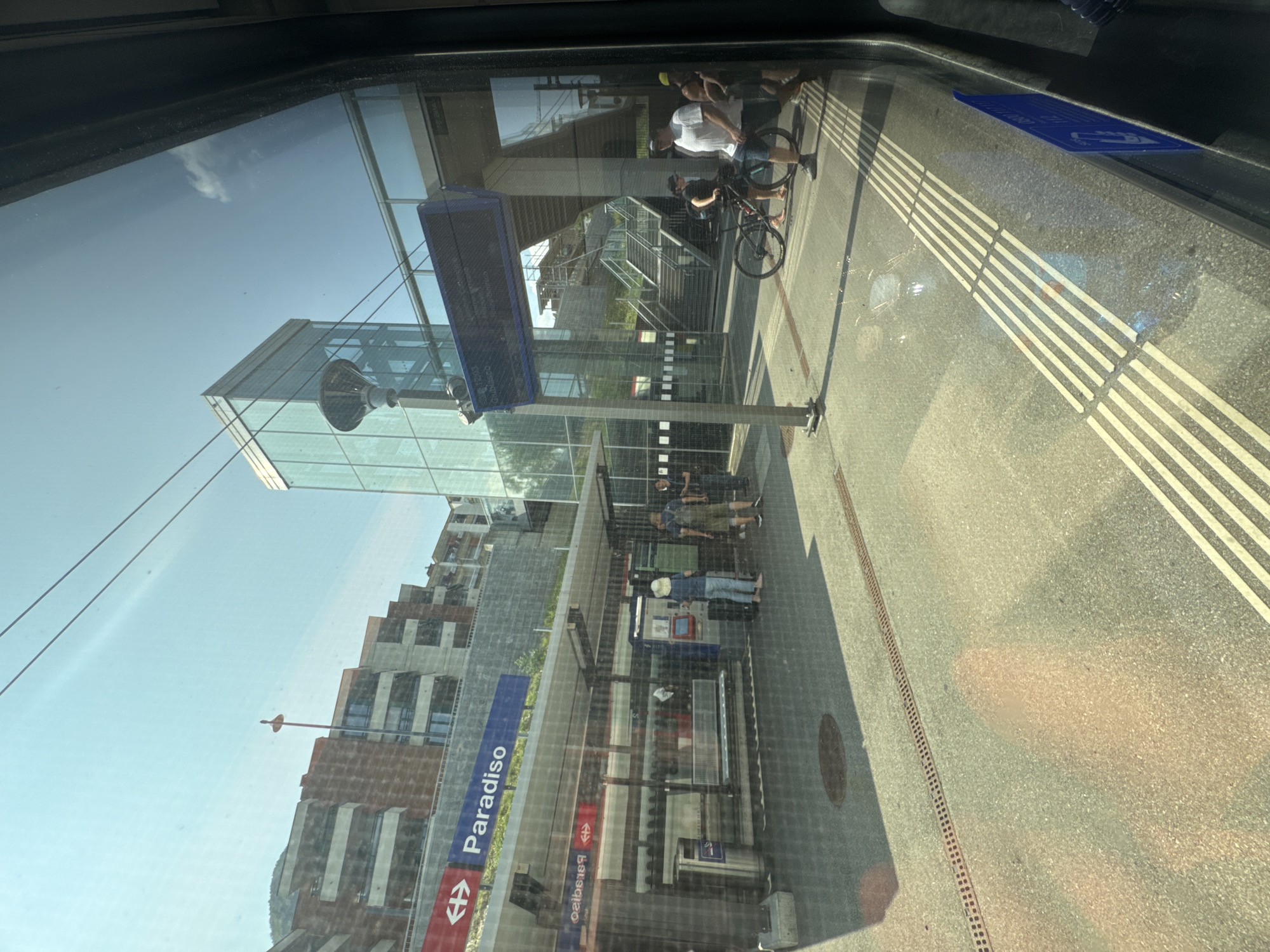 The image shows a view from inside a train or bus, looking out at the train station in Paradiso, Switzerland. A sign indicates "Paradiso" with the Swiss Federal Railways logo. The station features modern architecture with a glass building and an elevated walkway. Several people are visible on the platform, some with luggage. One person is near a ticket machine, while others are standing or walking. A bicycle is parked near a group of people. The platform has tactile paving for the visually impaired and a drainage grate. Sunlight casts shadows on the ground. The sky is clear and blue, with some overhead wires visible.