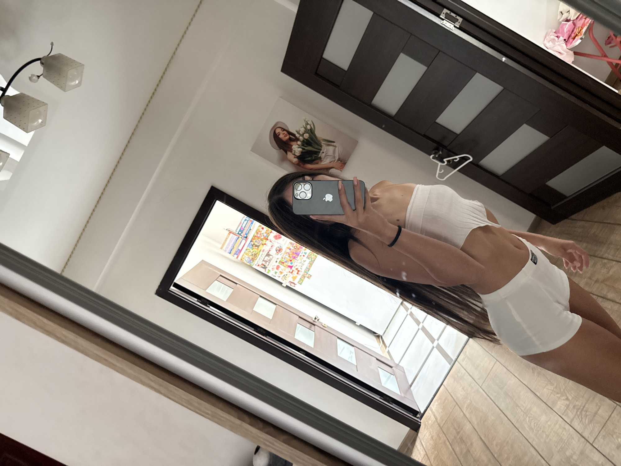 A young woman with long, dark hair is taking a mirror selfie in what appears to be a bedroom. She is wearing a white crop top and white shorts. The room has wooden floors and a dark brown door with frosted glass panels. There is a picture of a woman with tulips hanging on the wall, and various items are visible in the reflection of the mirror, including a doorway with tiled flooring and a ceiling light fixture. The scene is well-lit, likely by natural light. The location is not identifiable from the image alone.