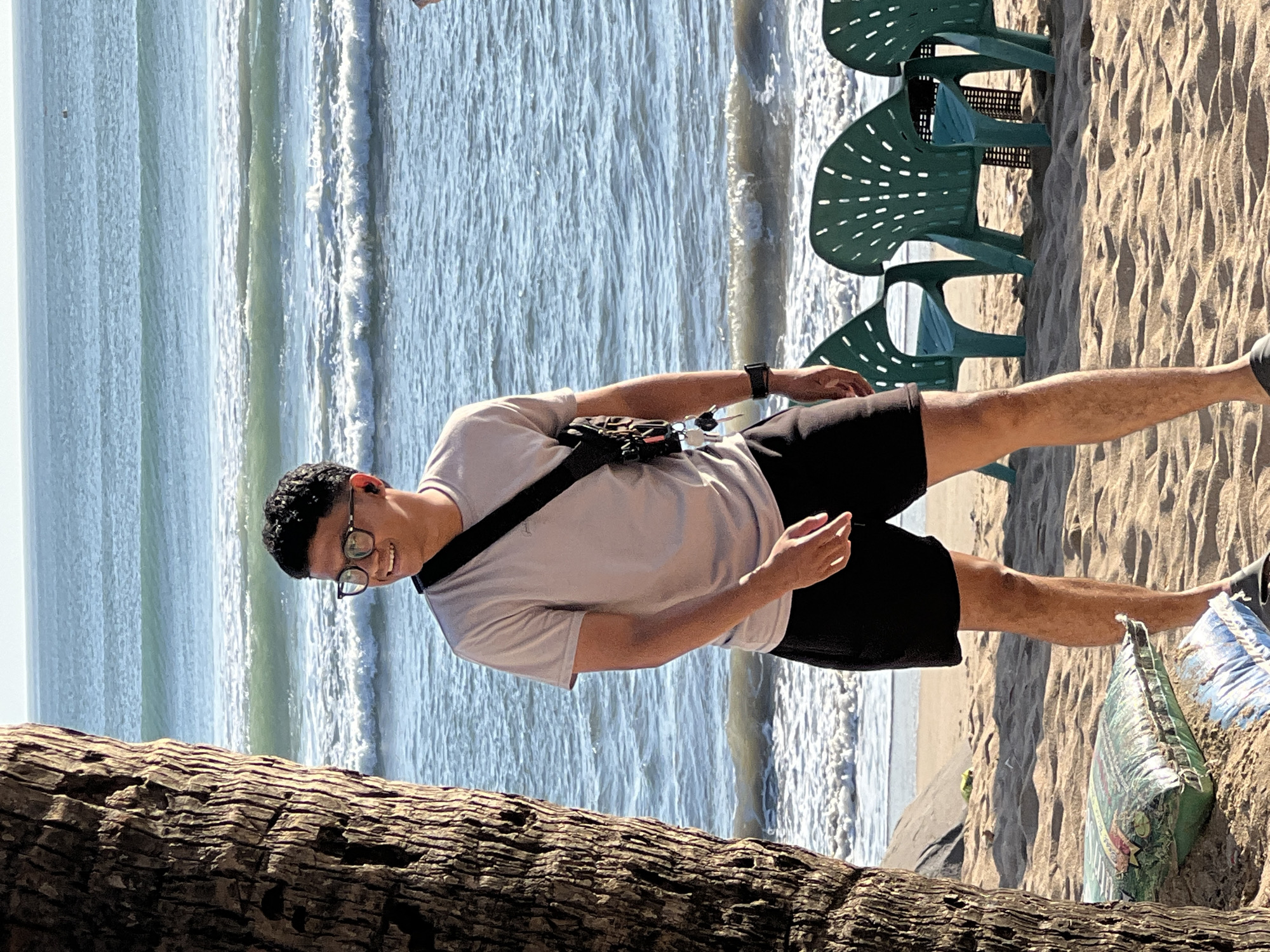 A man stands on a sandy beach in Kuta, Indonesia, with the ocean and waves in the background. He is wearing a light gray t-shirt, black shorts, glasses, and earbuds. A black sling bag is worn across his chest, with keys hanging from it. To his left, a portion of a palm tree trunk is visible. In the mid-ground, several green plastic chairs are arranged on the sand. There are also some bags on the sand in the foreground. The scene is illuminated by sunlight, suggesting it is daytime.