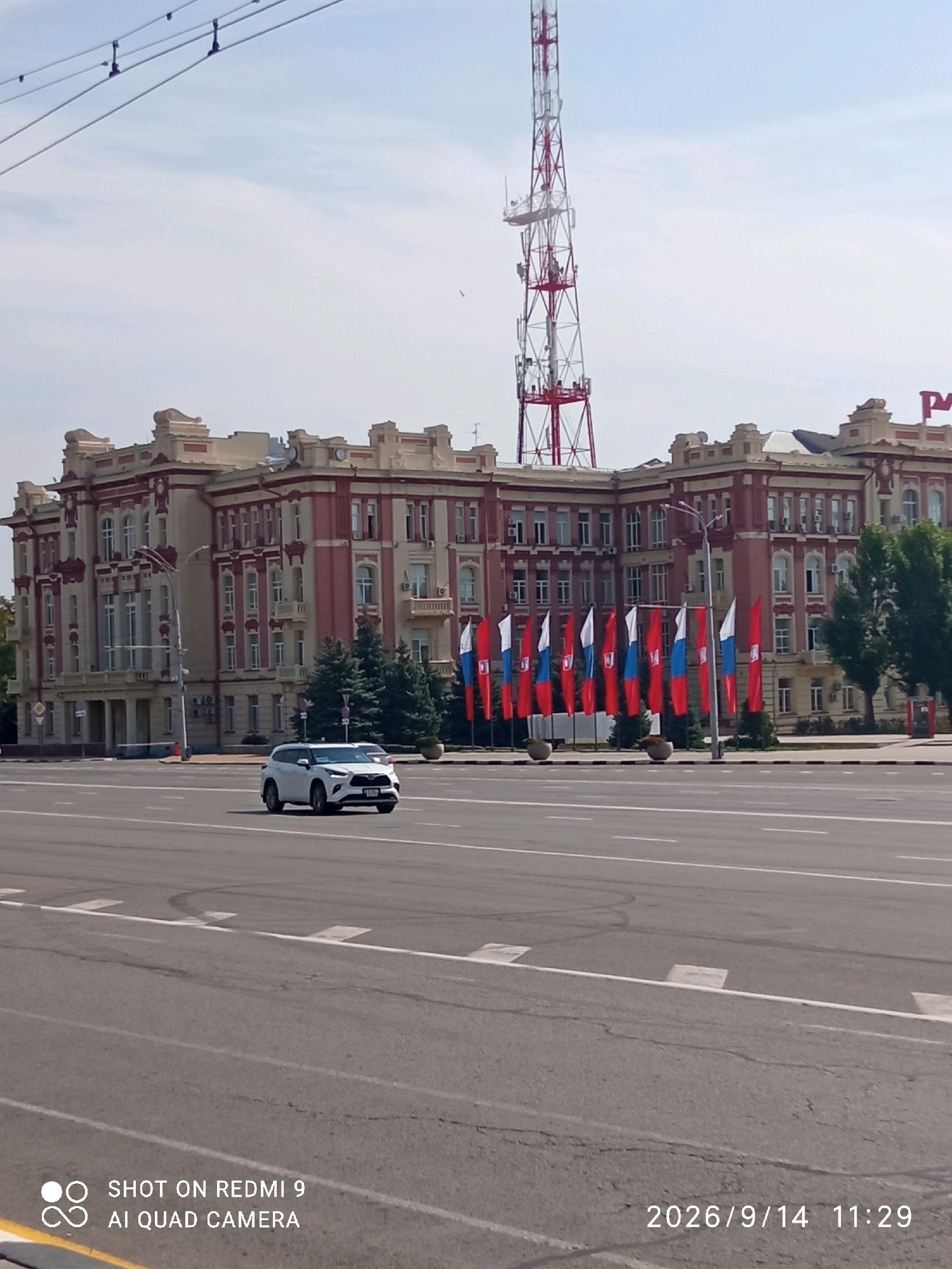 The image shows a wide, paved city street in Rostov-on-Don, Russia. In the background, a large, ornate building with red and beige detailing stands prominently. In front of the building, a line of Russian flags is displayed. To the right of the building, a tall, red and white radio tower is visible. A white SUV is driving on the street in the foreground, and electrical wires are strung across the top of the frame. The sky is partly cloudy. The bottom right corner of the image displays a timestamp of "2026/9/14 11:29" and the bottom left corner displays "SHOT ON REDMI 9 AI QUAD CAMERA".