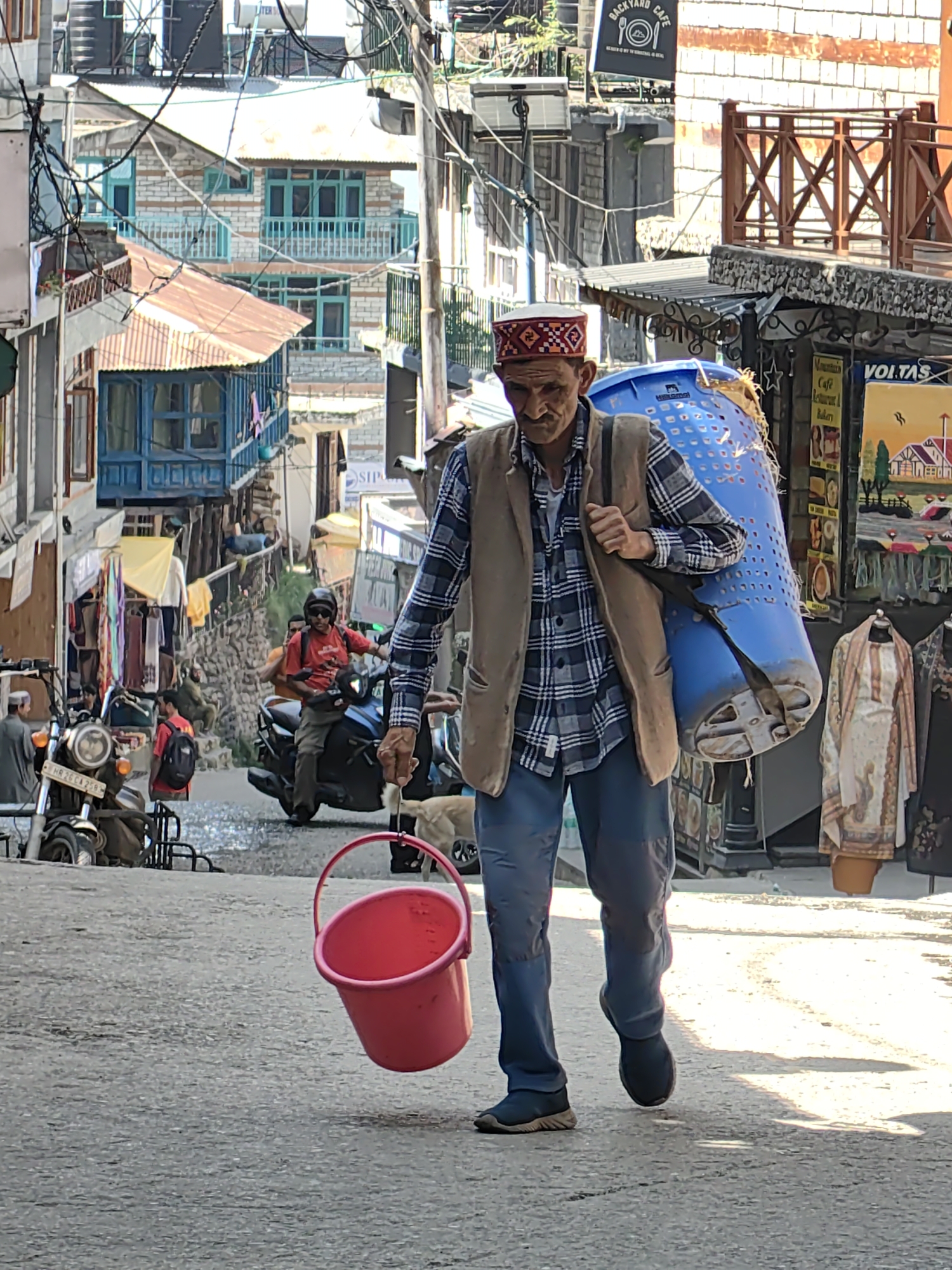 The image depicts a man walking downhill on a paved street in Vashisht, India. He is carrying a large blue plastic basket on his back, secured with a strap over his shoulder. In his right hand, he holds a pink bucket by its handle. The man is wearing a plaid shirt, a brown vest, blue trousers, and dark shoes. He has a cap on his head. Behind him, there is a dog and various shops and buildings lining the street, characteristic of a hilly town. Motorcycles are parked on the side of the street. The architecture suggests a traditional Indian mountain village. The overall scene captures a moment of daily life in Vashisht.
