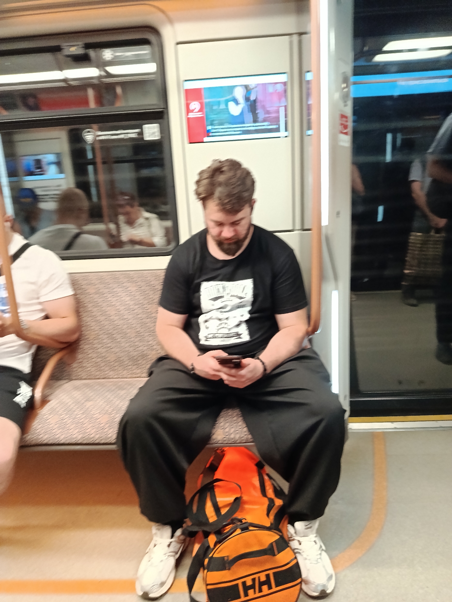 A man with a beard wearing a black t-shirt and black pants sits on a train, looking at his phone. He has a large orange duffel bag at his feet. Another person is visible sitting to his left. The train has beige seats and a window reflecting other passengers and the interior of the train. The floor of the train car is light gray with an orange stripe marking a safety zone. The overall environment suggests a public transportation setting.