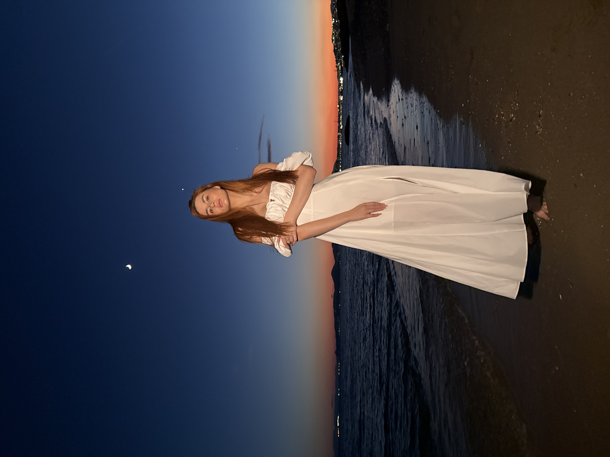 A woman with long, auburn hair stands on a sandy beach in Paleo Faliro, Greece, at dusk. She is wearing a white, off-the-shoulder maxi dress with a slit on the right side. Her arms are crossed in front of her. The sky behind her is a deep blue, transitioning to orange and pink near the horizon, indicating the sunset. A crescent moon is visible in the upper left portion of the sky. The sea is to her left, with gentle waves lapping the shore. In the distance, across the water, a city skyline with lights is visible, likely part of the Athens metropolitan area. The beach itself is dark and sandy, with some small shells and debris scattered on the surface. The overall scene conveys a serene and romantic atmosphere.