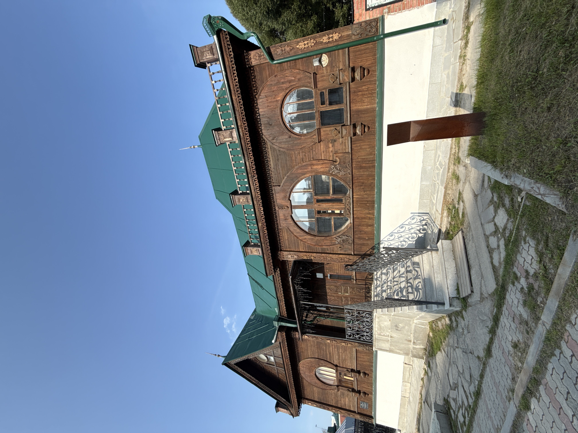 This image depicts the House of Crafts (also known as the "House of Merchants" or "Sverdlovsk Regional Museum of Local Lore") located at 11 Karl Liebknecht Street in Yekaterinburg, Russia. The building is a two-story wooden structure with a prominent green metal roof. It features intricate wood carvings along the eaves and around the windows. A distinctive circular window is centrally placed on the facade. A set of stone steps with ornate black metal railings leads to the entrance. The building is situated on a paved area with flagstone and brick pathways, and a patch of grass with a single, tall, rusty metal object is present in the foreground. The sky is clear and blue, suggesting a daytime setting under fair weather conditions. There are no people visible in the image.