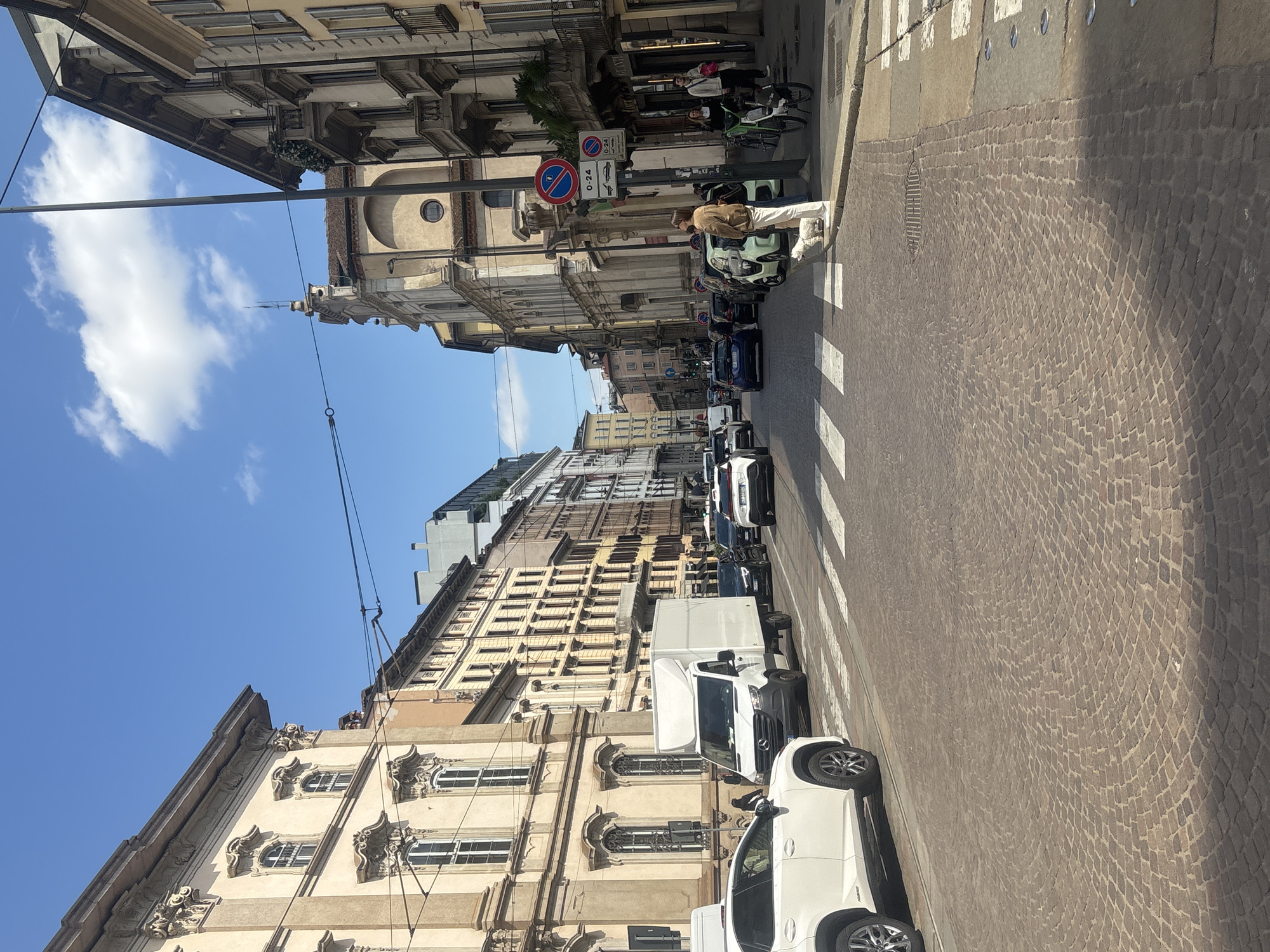 This image presents a vibrant street scene in Milan, Italy, captured on a clear, sunny day, likely during the late morning or early afternoon. The perspective looks down a historic street flanked by tall, architecturally rich buildings, featuring multiple stories, ornate window dressings, and decorative cornices characteristic of European design.
The foreground is dominated by a cobblestone street, intricately laid with small, dark-hued stones, and crossed by a pedestrian zebra crossing with distinct white stripes. Several vehicles are parked along the right side of the street, including a white SUV, a white delivery van, and various other cars.
In the midground, a person wearing a tan jacket and white pants stands on the pedestrian crossing, facing to the left. A light green compact car is parked nearby. Further down the street, two individuals are visible near bicycles, with one seemingly seated on a bike and the other standing alongside it. On the right side, a street sign is clearly visible, showing a blue circular sign with a red diagonal line indicating "No Stopping" or "No Parking." Below it, a white rectangular sign displays "0-24," likely denoting the hours of the restriction, and a smaller sign with a vehicle icon is also present.
The background features more of the city's architecture, blending into the distance. The sky above is a brilliant, clear blue with a few scattered white cumulus clouds. Overhead power lines crisscross the upper portion of the frame, hinting at the city's infrastructure. The overall scene conveys the bustling yet charming atmosphere of an Italian urban environment.