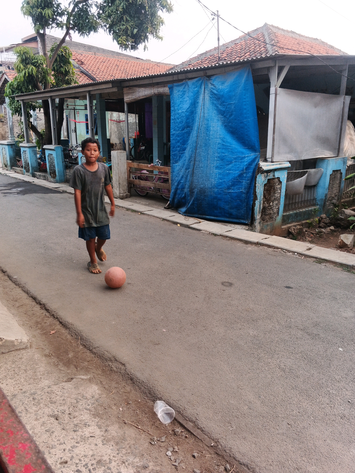 A young boy, appearing to be of Indonesian descent and wearing a dark t-shirt and blue shorts, is walking on a paved road in Kadu, Indonesia. He is barefoot, wearing sandals. In front of him on the road is a pink ball. The boy is looking towards the camera with a neutral expression. To his left, a blue fence with decorative posts runs along the side of the road, leading to a building with a red tiled roof. A large blue tarp is draped over a section of the building, and part of the building's facade is painted blue and appears to be crumbling. The environment suggests a residential area, with some trees visible and power lines overhead. Debris, including a discarded plastic cup, is scattered on the side of the road. The lighting indicates it is daytime.