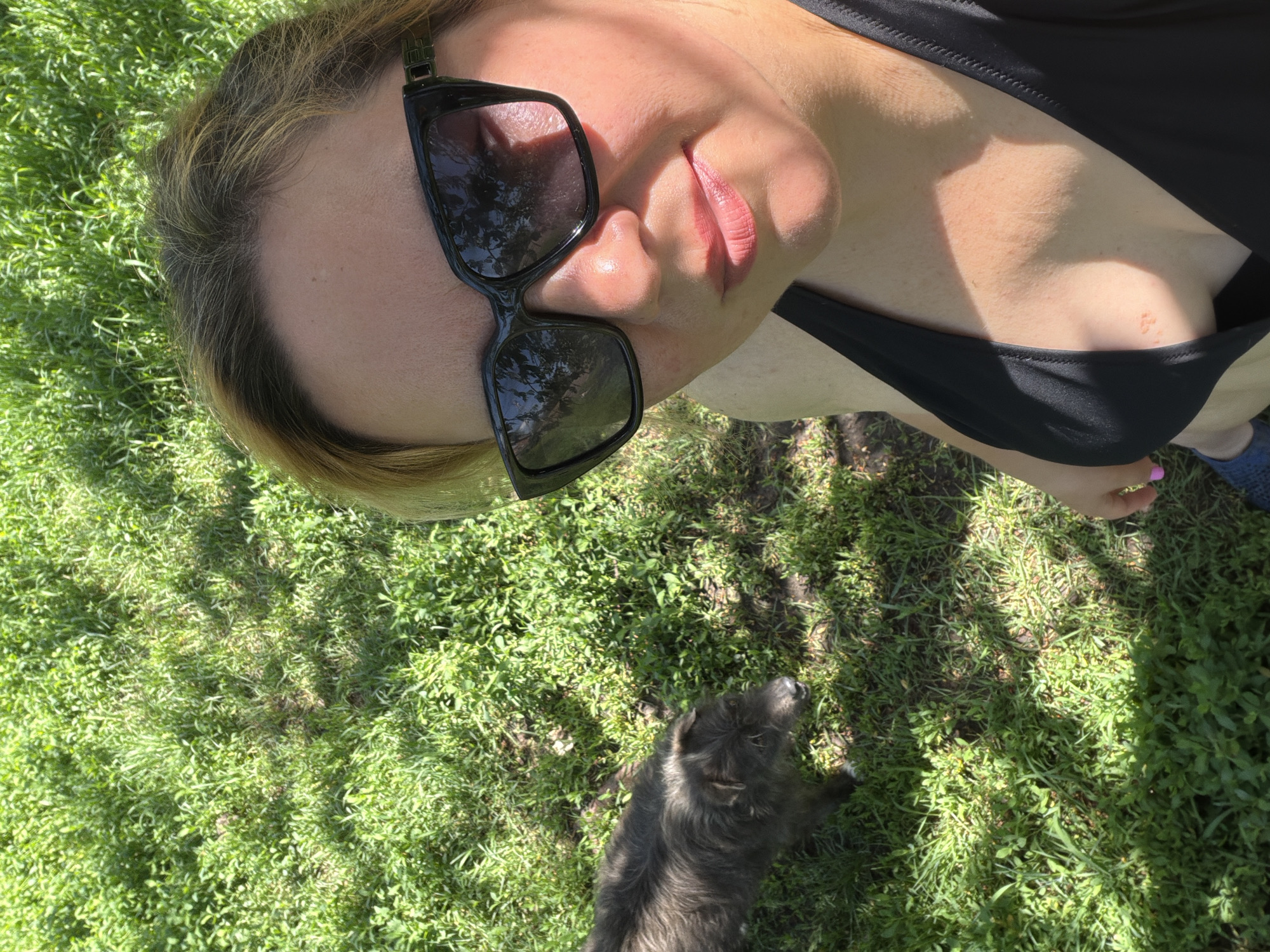 A woman wearing sunglasses and a black top is lying on green grass. A small gray dog is standing nearby. The location is outdoors, likely in a grassy area. No context indicates Russia.
