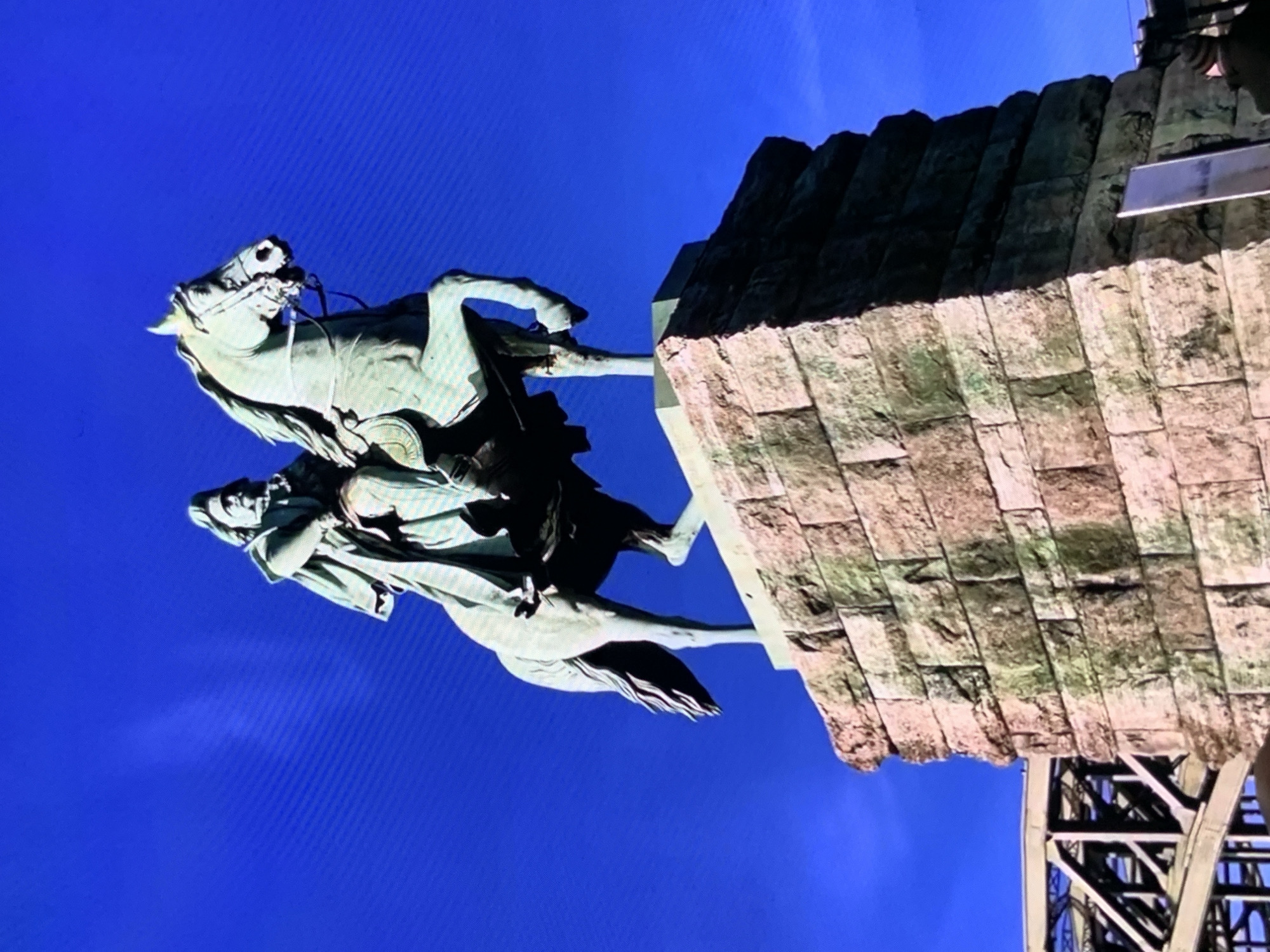 An equestrian statue of General Alexander Suvorov stands atop a stone pedestal against a clear blue sky. The statue depicts Suvorov in military attire, seated on a horse that is rearing on its hind legs. The horse and rider are made of bronze, exhibiting a greenish patina. The pedestal is constructed of large, rough-hewn stone blocks, with visible signs of weathering and some moss or algae growth. To the left of the pedestal, a portion of a metal bridge structure is visible. The overall scene is illuminated by daylight.