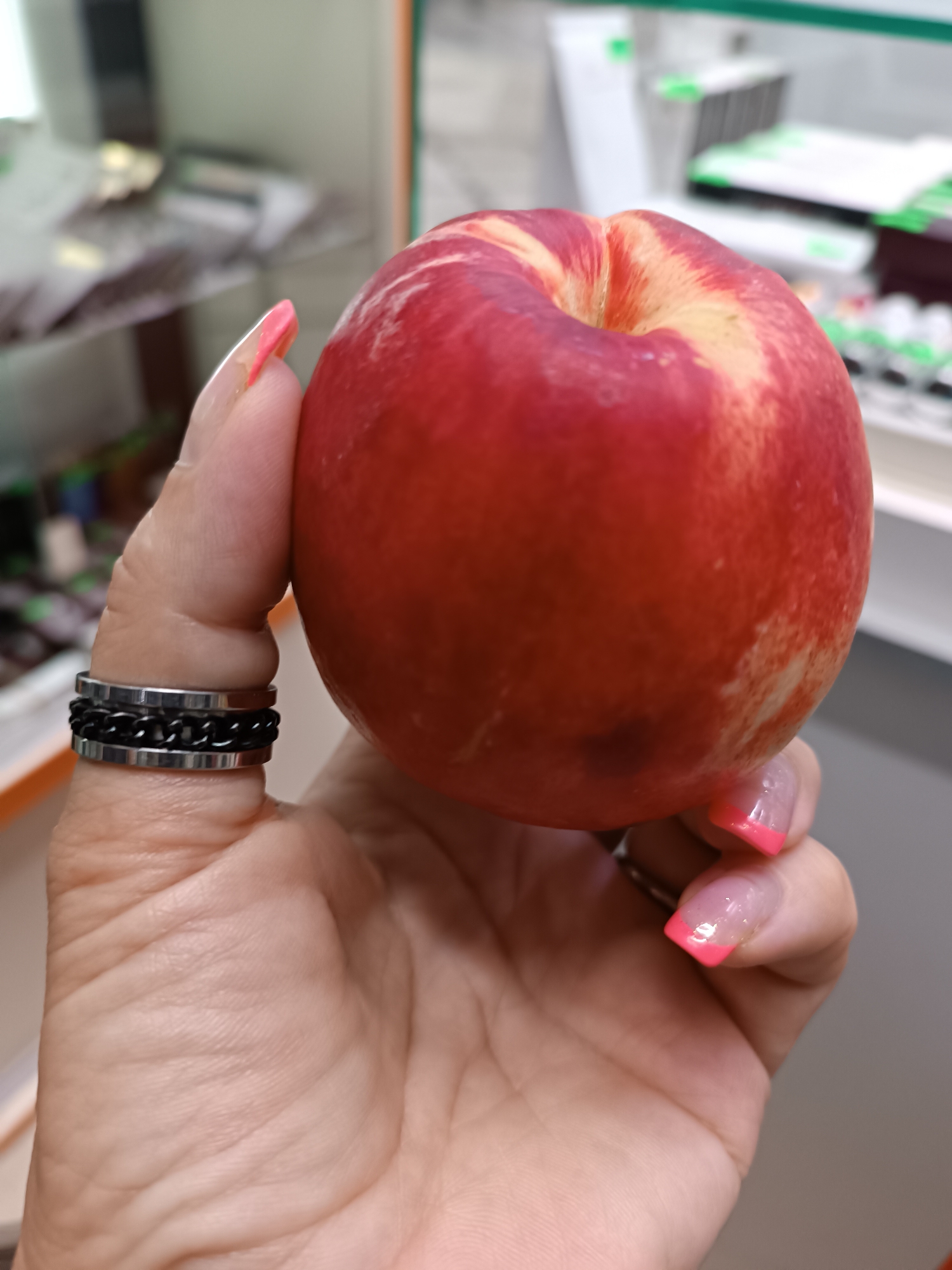 A close-up shot shows a person's hand holding a ripe nectarine. The hand has bright pink French manicured nails and is wearing a silver ring with a black chain-link design. The nectarine is predominantly red with streaks of yellow and some darker, bruised spots. The background appears to be an indoor setting, possibly a grocery store or market, with shelves and products visible but out of focus. The overall lighting is bright and even.