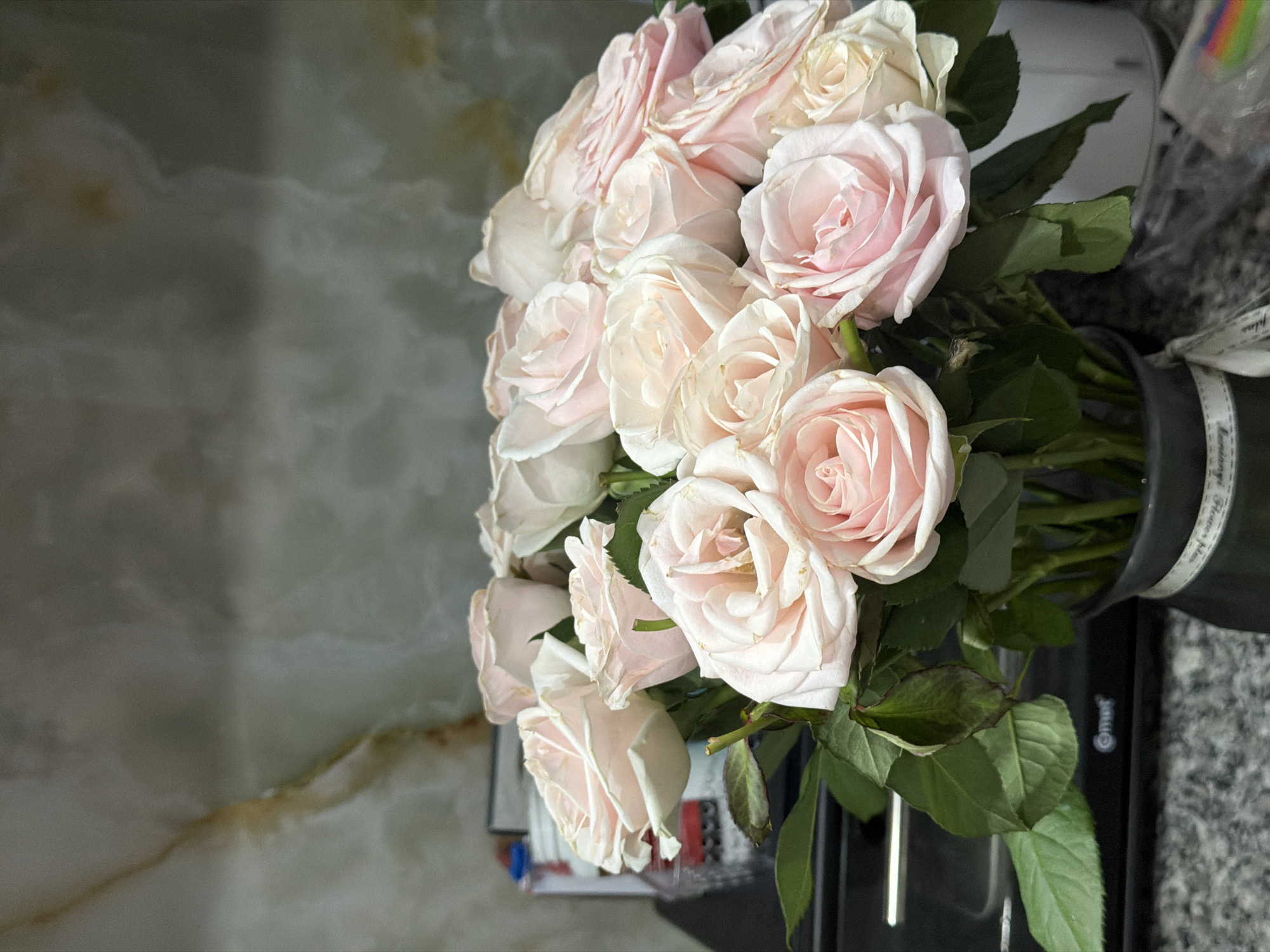 A still life image shows a bouquet of light pink roses in a dark-colored vase, placed indoors on a dark surface that is on top of a light-colored marble surface. The roses are in full bloom with petals of varying shades of light pink. The green leaves of the roses are visible around the blooms. The vase is partially obscured, but its cylindrical shape and dark color are apparent. The indoor setting is suggested by the surfaces beneath and behind the floral arrangement. The location is Nha Trang Ward, Vietnam.