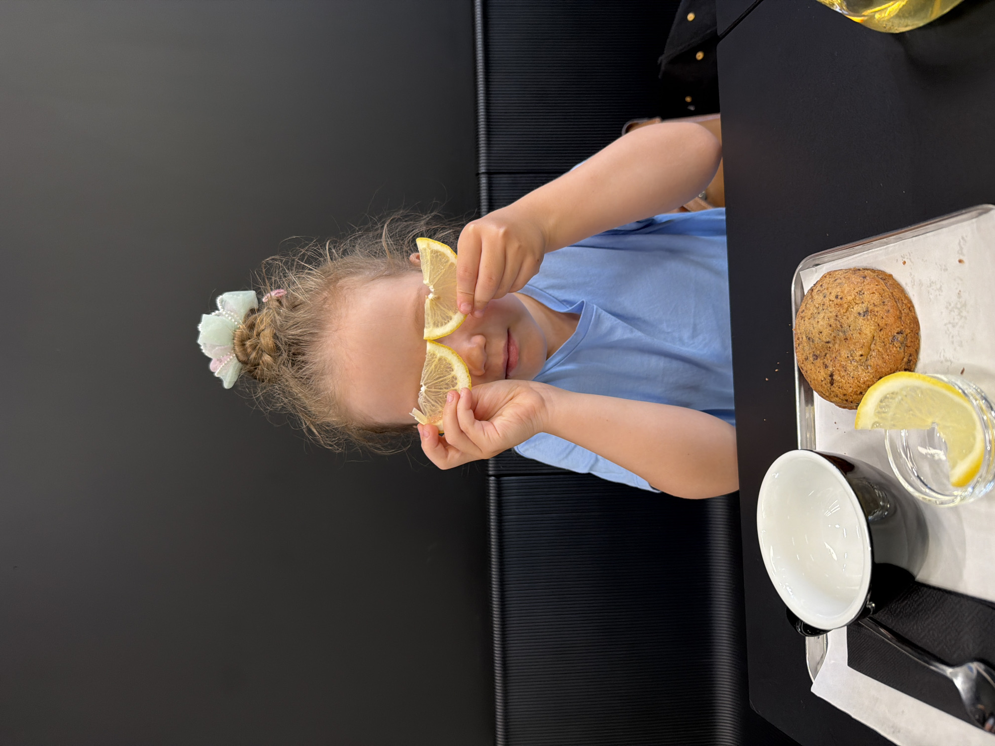 A young girl with blonde hair tied in a bun with a pastel bow is sitting at a black table, holding two lemon slices over her eyes as if they were glasses. She is wearing a light blue t-shirt. In front of her on a silver tray is a black cup, a small glass dish with a lemon slice, and a round cookie or pastry. The background is a dark textured wall. The image captures a moment of playful interaction with food in what appears to be a cafe or restaurant.