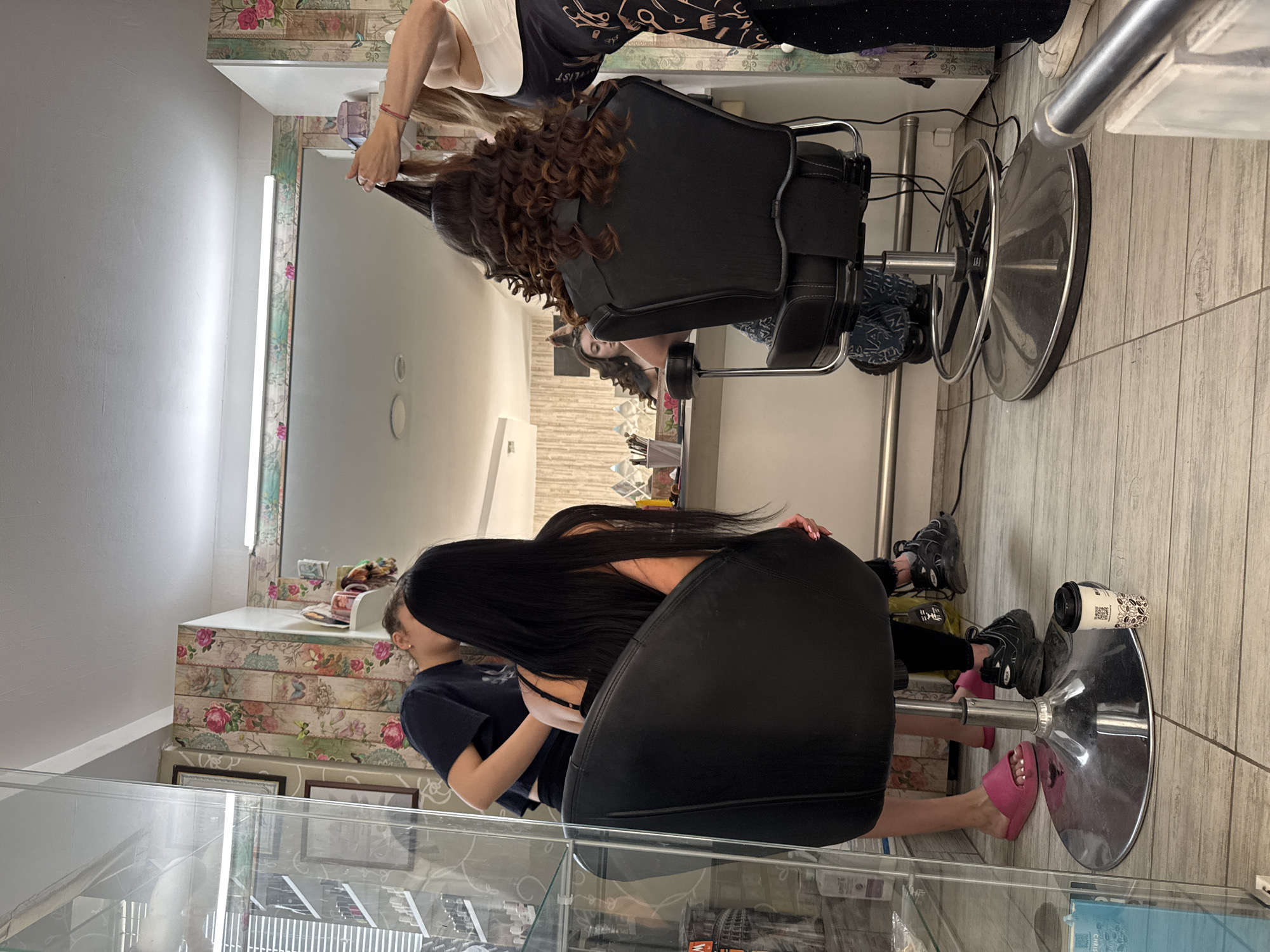 A salon in Dolni Chiflik, Bulgaria has two women in it. A brunette with long, straight black hair is in a black salon chair; a woman with shoulder-length curly auburn hair is in a similar black chair. A stylist in a white shirt is working on the auburn-haired woman. The salon has a large mirror. Below a glass display case, there are objects on the floor. The floor is light wood; one of the women is wearing pink slippers, and a coffee cup is on the floor.