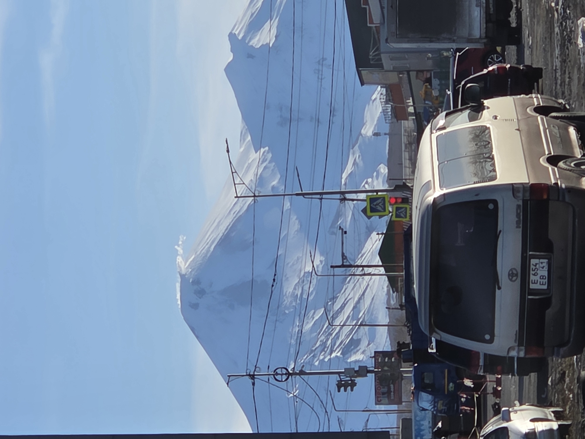 The image shows a street scene in an urban environment. A large, snow-covered mountain dominates the background. In the foreground, there is a white van with the license plate "E654 EB 41". A traffic light is visible with a red light illuminated. Overhead power lines are also visible in the middle ground. The sky is clear blue.