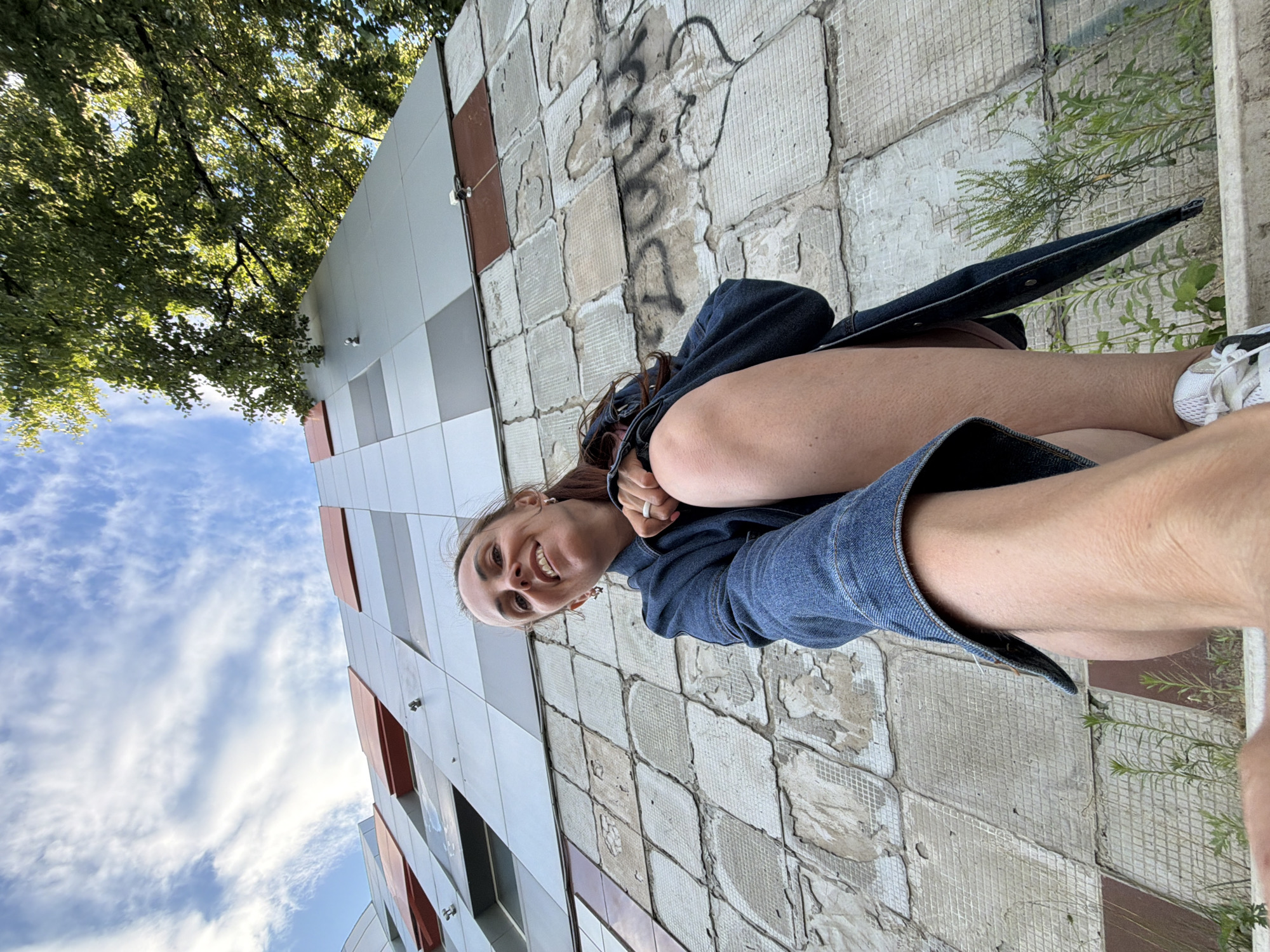 A woman with brown hair is crouching down in front of a building with a textured, tiled facade and a modern upper section with red and grey panels. She is wearing a denim jacket and white sneakers. The building appears to be in an urban setting, with trees visible in the background under a cloudy sky. Graffiti is visible on the lower section of the building's facade. The photo is taken from a low angle, looking up at the woman and the building. It is not possible to confirm the location in Birobidzhan, Russia, based on the image alone.