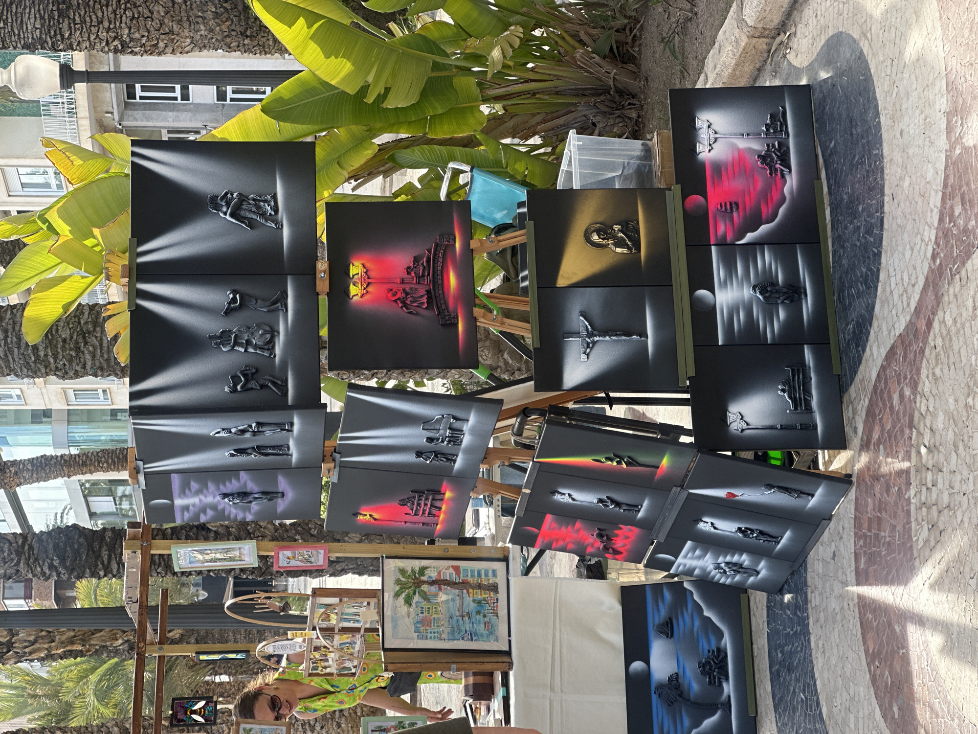 The image captures an outdoor art stall, likely located on a promenade or street in Alicante/Alacant, Spain, identifiable by the distinctive mosaic pavement and abundant palm trees. Numerous art pieces are displayed on wooden easels and racks under bright daylight, casting clear shadows.
The primary subjects are the art pieces, predominantly spray paint art on dark canvases. These artworks feature vibrant spray-painted atmospheric effects in colors like red, white, and blue, often highlighting silhouetted figures or small, dark, three-dimensional elements attached to the canvas. Common motifs include figures of musicians, dancers, religious symbols such as crucifixes, mythological creatures, and abstract shapes, frequently accompanied by moon or sun-like spheres. A few traditional watercolor-style paintings are also visible, including one depicting a coastal scene with a palm tree and buildings.
In the lower left foreground, a woman, presumably the artist or a vendor, is partially visible, looking down, suggesting she is attending to the stall or merchandise. Behind the art display, large green leaves resembling banana plants and sturdy palm tree trunks are prominent, with glimpses of modern buildings and a lamppost further in the background. Clear plastic containers are also visible behind some of the art.
The activity taking place is an outdoor art sale or display. The time of day is bright and sunny, indicated by the strong illumination and crisp shadows. The mosaic pavement, with its intricate patterns of black, white, grey, and red-brown, serves as a strong location cue for a Spanish coastal city. There is no clearly visible text on any of the art pieces or signs. The overall atmosphere appears relaxed and commercial, typical of a tourist-oriented area.
