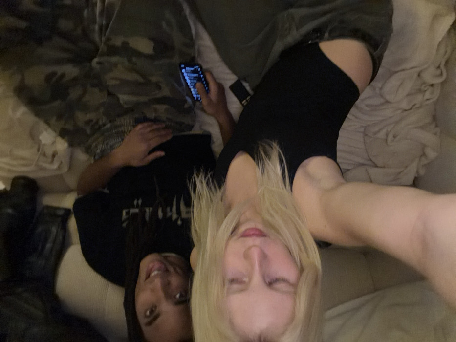 Two women lounge on a sofa or bed. The woman on the left has long, dark hair and is smiling. She wears a black t-shirt. The woman on the right has long, blonde hair and her eyes are closed. She is wearing a black bodysuit. A phone is visible in the hands of the woman on the left. One woman wears camouflage print pants and the other a pair of light green pants. Blankets and additional items are also visible on the surface.