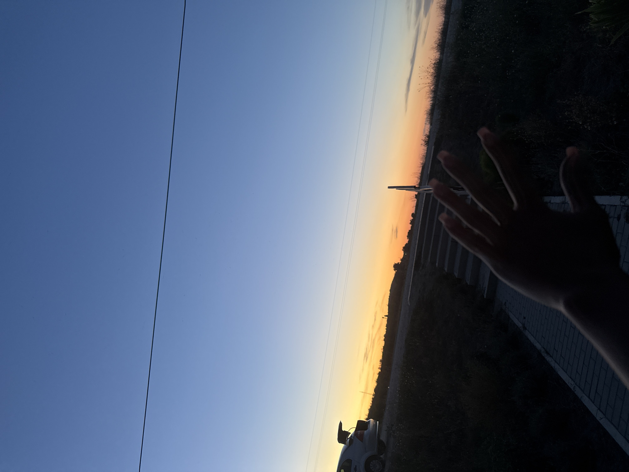 A silhouette of a hand with outstretched fingers is in the foreground of an image taken at dusk. Behind the hand, a paved path leads to a set of concrete stairs. To the left of the stairs, a car with its trunk open is parked on the side of a road. The horizon features a band of warm, colorful light from the setting sun, transitioning to a clear, pale blue sky. Power lines are visible horizontally across the sky. The location is Cimișlia, Moldova.