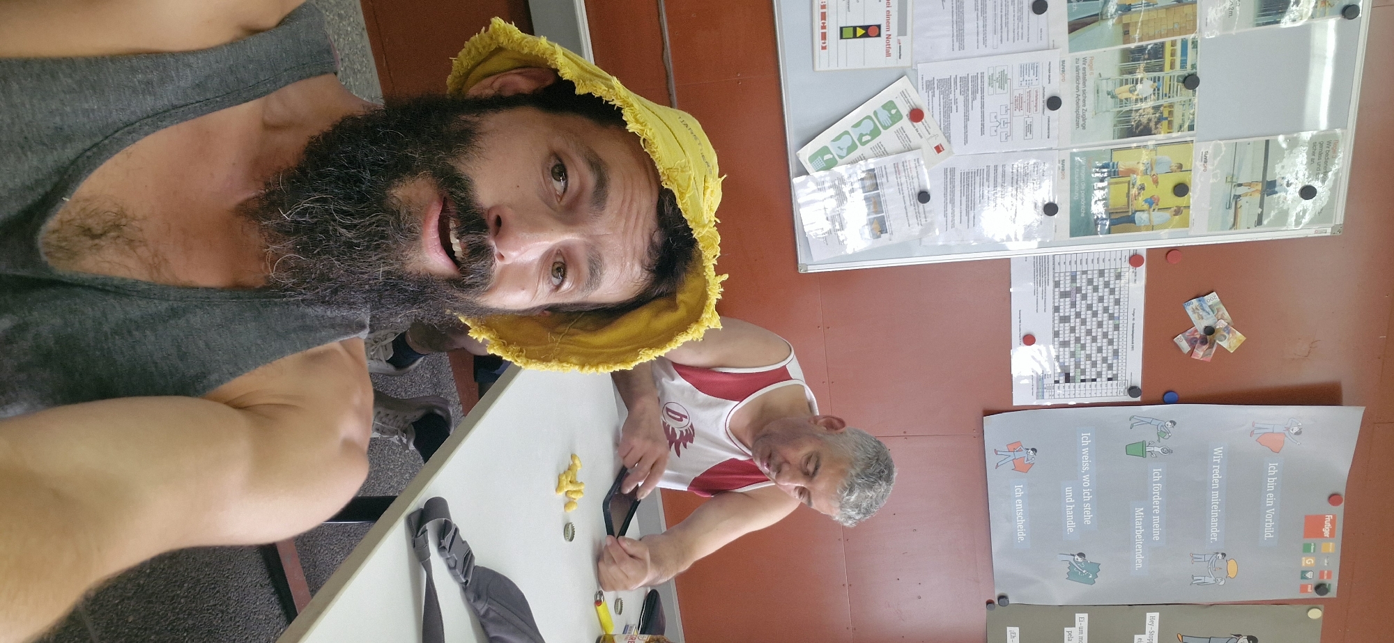 In Uetendorf, Switzerland, two men are seated at a white table. The man in the foreground, with a prominent beard and a yellow bucket hat, takes a selfie. He wears a grey tank top. The man in the background, with grey hair, sports a white tank top with red trim. He appears to be using a smartphone. On the table, between the two men, are scattered yellow corn snacks, a mobile phone, a yellow lighter, and a bottle cap. The environment includes a bulletin board covered with various papers and posters on the left and a large poster with German text and illustrations on the right. The wall behind the men is a warm, reddish-brown color. The floor is carpeted with a dark, speckled pattern.