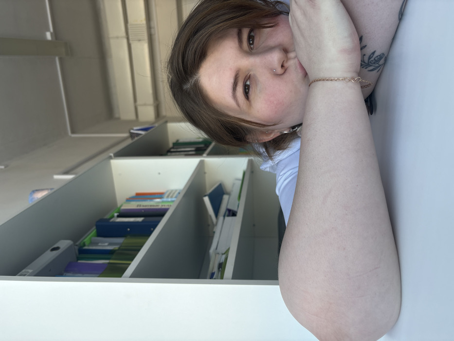 The image is a portrait-style photograph of a young person taken indoors, rotated 90 degrees counter-clockwise.
The central subject is a young individual with medium-brown, shoulder-length hair and fair skin. They have light-colored eyes and a small silver stud piercing on their left nostril, as well as multiple piercings visible on their left ear. Their cheeks show a natural rosy blush. They are wearing a light-colored (possibly white or light blue) short-sleeved top. On their right wrist, a delicate gold-colored chain bracelet is visible. A tattoo, which appears to be a floral or organic design, is visible on their upper left arm, near the elbow.
The person is resting their head on their right arm, which is placed on a smooth, light-colored surface, likely a desk or table. Their left arm is extended forward, partially obscuring their body. They are looking directly at the camera with a relaxed, calm expression, their lips slightly parted in a subtle, thoughtful pose.
The background primarily consists of a white shelving unit or bookcase with multiple compartments, filled with various folders and books. These items are mostly in shades of blue, green, and orange, and many appear to be ring binders. Above the shelving unit, in the upper portion of the background, architectural details such as light-colored ceiling beams or ventilation ducts are visible, suggesting an institutional or office setting.
The lighting is bright and natural, indicating the photo was taken during the daytime. Most of the text on the book and folder spines in the background is unreadable due to the distance and resolution, but some horizontal lines of text are discernible. One particular green binder appears to have white text on its spine, and another spine seems to feature Cyrillic script, though specific words cannot be clearly identified.
The overall impression is a candid, serene moment, possibly a break from studying or work, captured in a brightly lit interior space.