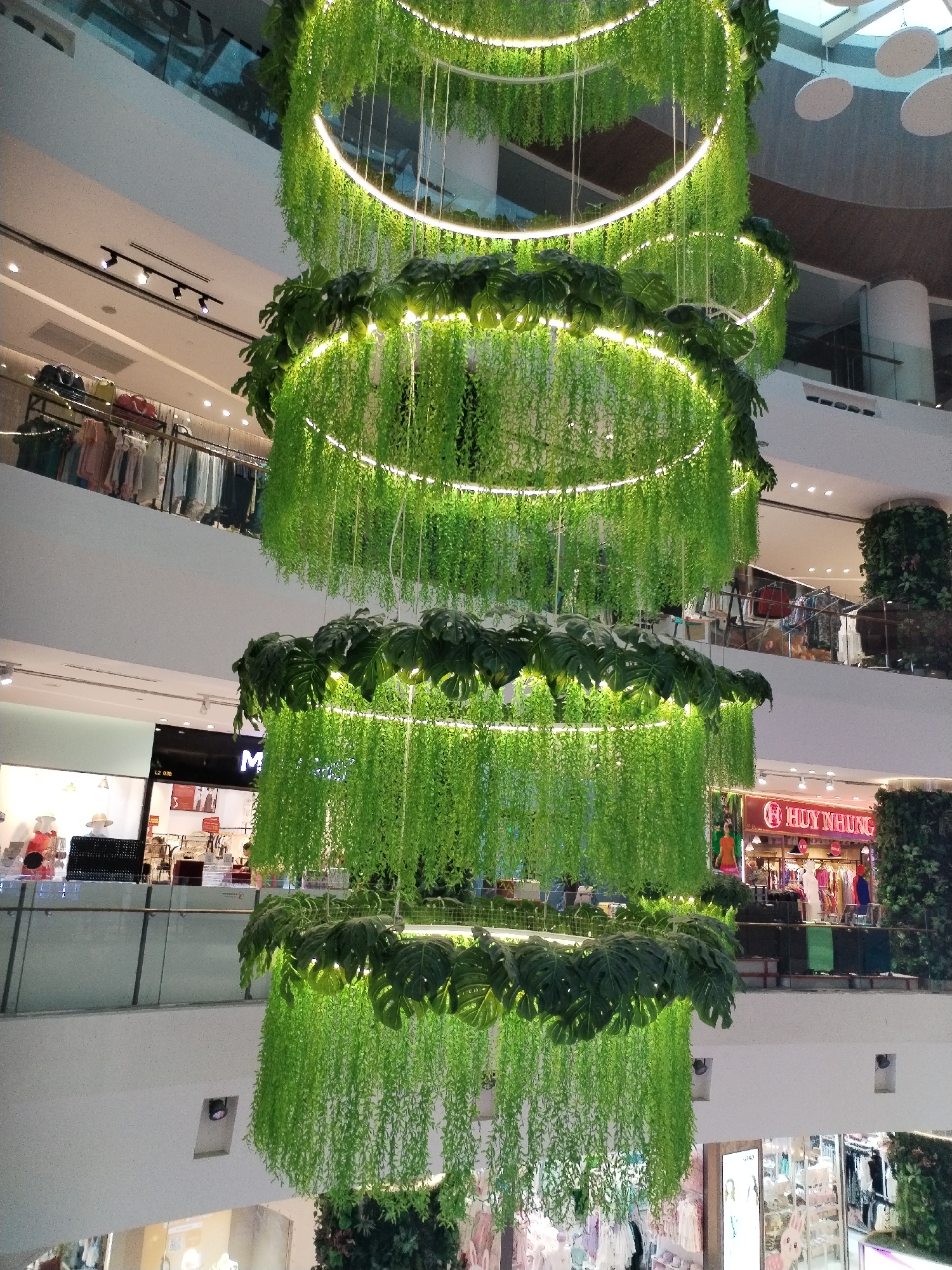 The image shows the interior of a multi-level shopping mall in Nha Trang Ward, Vietnam. The central focus is a large, tiered hanging decoration made of artificial green plants and large monstera leaves, illuminated by ring lights. The decoration cascades down through several floors of the mall.
In the background, visible on different levels, are various retail stores. One store prominently displays the name "HUY NHUNG" in red lettering on a gold sign. Racks of clothing are visible in store windows and on display. People are seen browsing in some of the stores and walking along the mall's walkways. The mall architecture features white walls and railings, with some areas showing exposed ceilings with lighting fixtures and air conditioning vents. On the upper levels, more clothing displays and store signage are visible. The overall environment suggests a brightly lit, modern retail space.