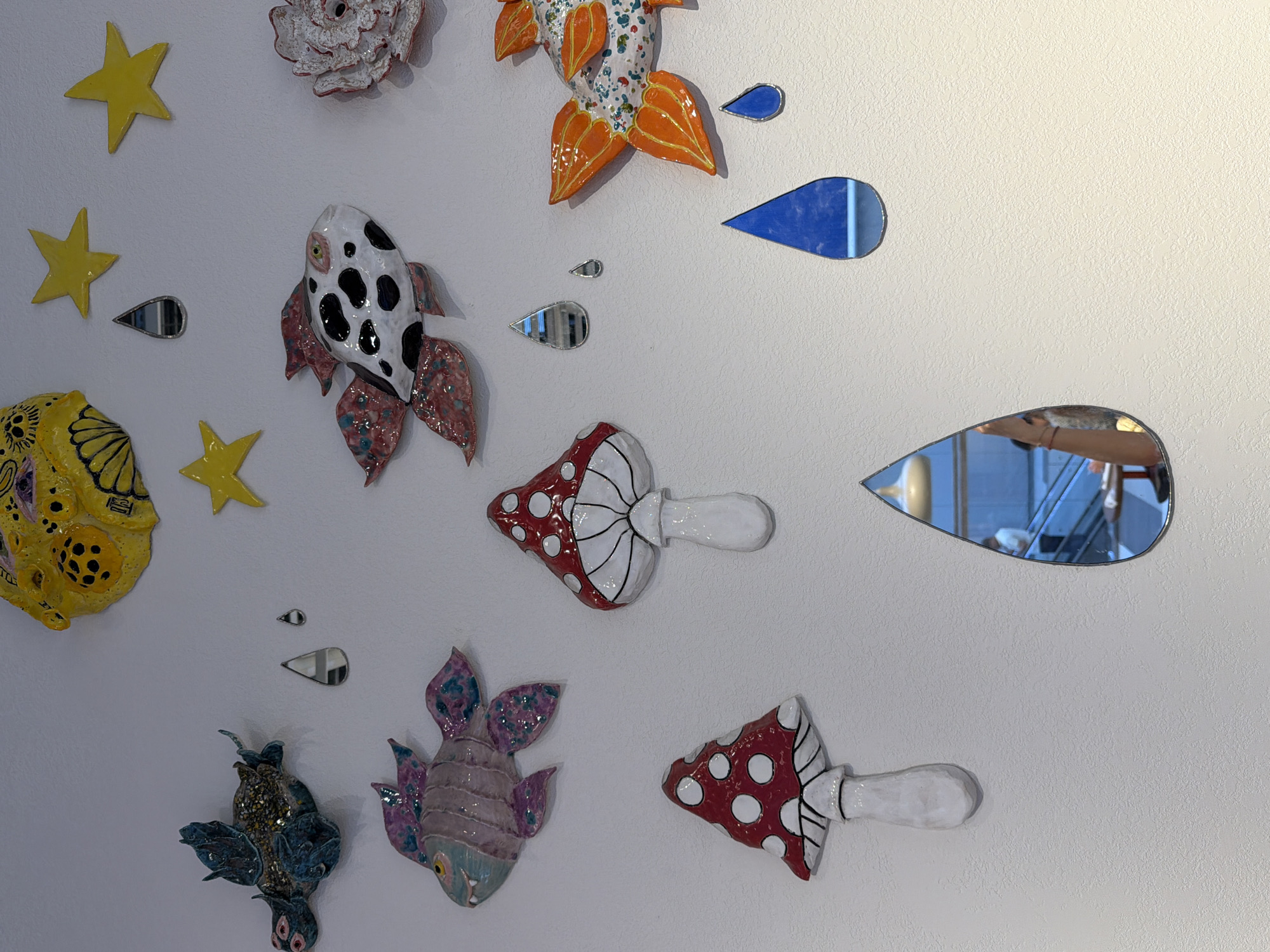 The image depicts a wall adorned with various ceramic art pieces and small mirrored decorations in the shape of water droplets. The ceramic art includes several stylized fish with colorful scales, two mushrooms with red caps and white polka dots, a larger yellow object with circular patterns, and a pinkish-white flower-like object. Yellow star shapes are also attached to the wall. The mirrored droplets reflect parts of the environment, including what appears to be interior architecture with stairs and a person visible in one of the larger droplets. The wall itself is textured and painted white, with a warm light source illuminating the scene from the right side of the frame, casting a soft shadow. The overall impression is of a whimsical and artistic display.
It is not possible to determine the specific location in the city of Yekaterinburg, Russia from the provided image.