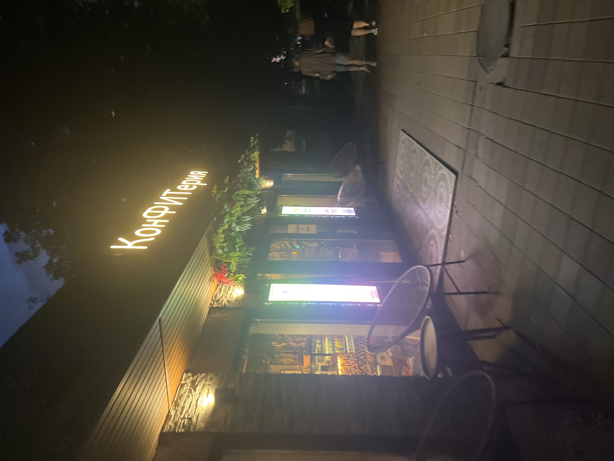 The image shows the exterior of a confectionery shop at night in Krasnodar, Russia. A neon sign above the entrance reads "Кондитерская" (Confectionery). The shop has a brick facade and large glass windows, revealing displays inside. Outside the shop, there are several outdoor seating areas with chairs and tables. Two people are walking on the sidewalk in front of the shop. The lighting is dim, with warm light emanating from the shop's interior and the neon sign. The overall scene suggests a pleasant evening at a local establishment.