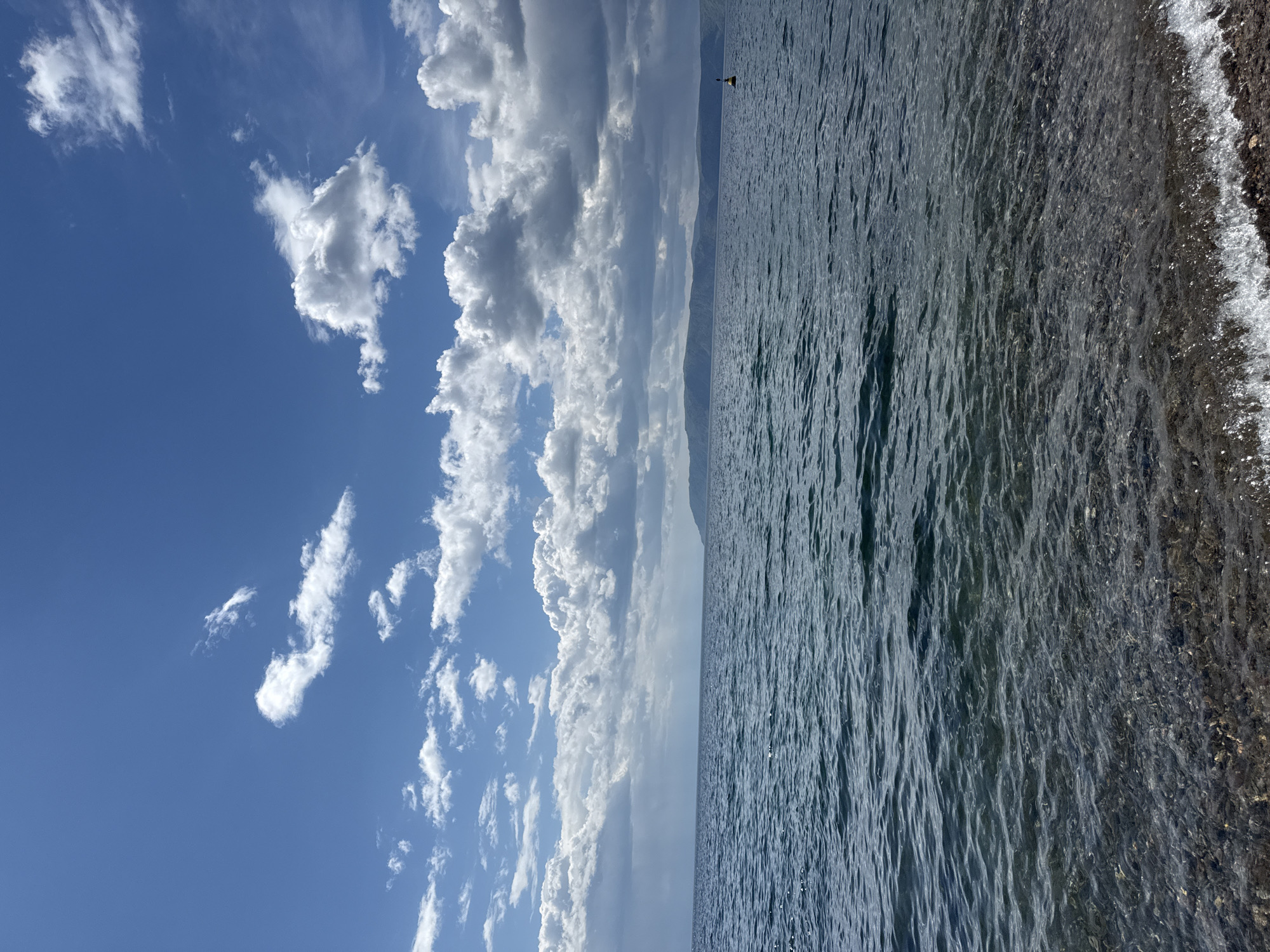 The image depicts a landscape divided between a vibrant blue sky with scattered cumulus clouds and a body of water, likely a lake or sea. The clouds range in size and density, some appearing wispy and others more substantial. A thin strip of land or mountains is visible in the distant center of the frame, providing a horizon line. The water surface is textured with ripples and waves, transitioning from a darker hue in the center to lighter tones near the shoreline. The shoreline consists of small rocks or pebbles, suggesting a natural beach.
The location is plausibly Listvyanka, Russia, given the typical landscape features of Lake Baikal: the combination of clear blue skies with distinctive cloud formations, the vast expanse of water with a distant shoreline or mountain range, and the rocky or pebbled beaches that characterize the lake's edge. These elements align with the known geographical characteristics of Listvyanka and the surrounding Lake Baikal region.
