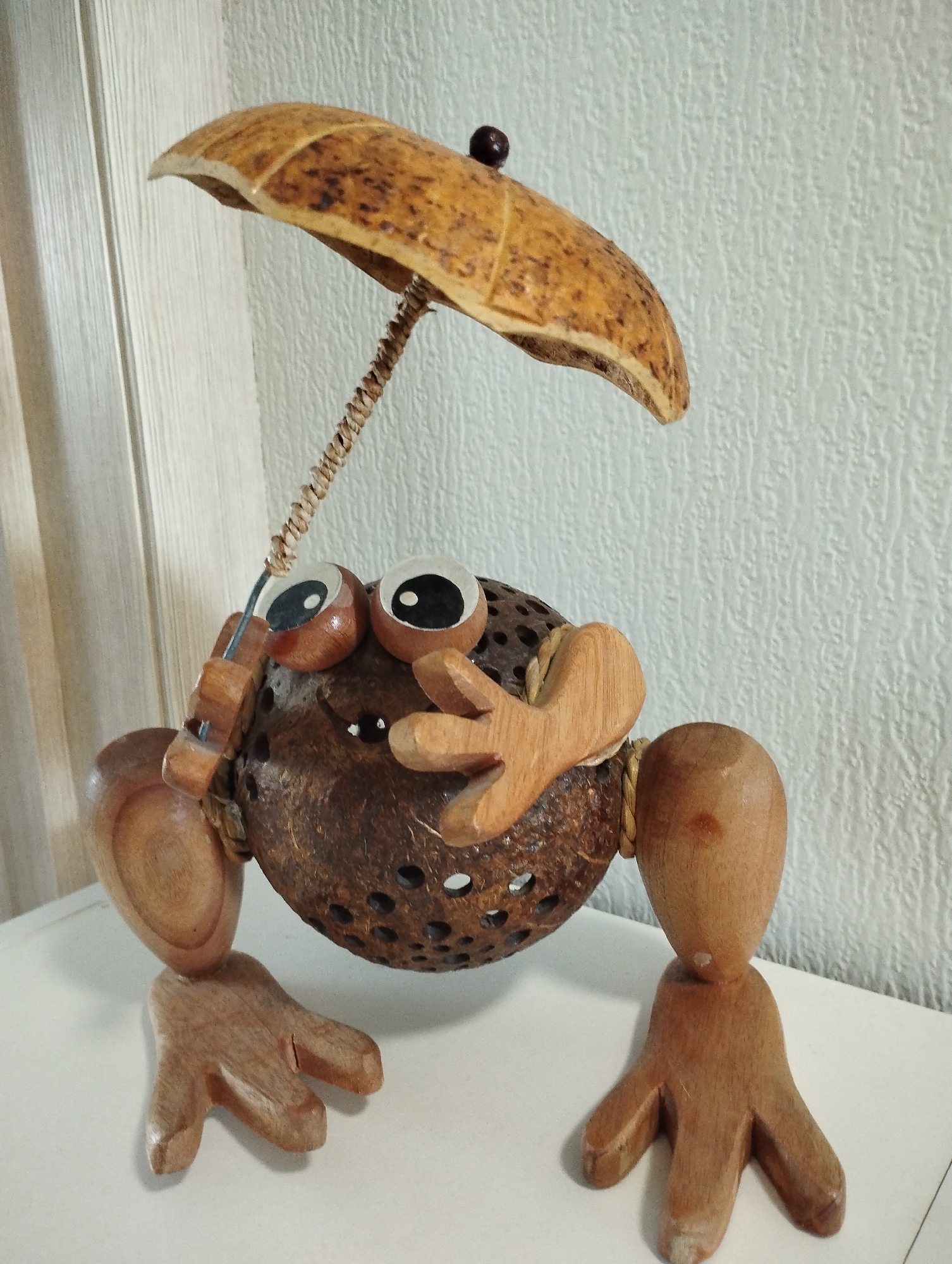 A wooden figure of a frog-like creature stands on a white surface in front of a textured white wall. The creature has a round body made from a coconut shell with holes drilled in it. Its eyes are round and white with black pupils, and it has wooden hands, feet, and arms attached with string. The frog-like creature holds a small umbrella made from a piece of wood. The background shows a white surface and a textured white wall. There are no people or other subjects visible in the image. The scene is brightly lit. The location of the image is specified as Sadovyy, Russia.