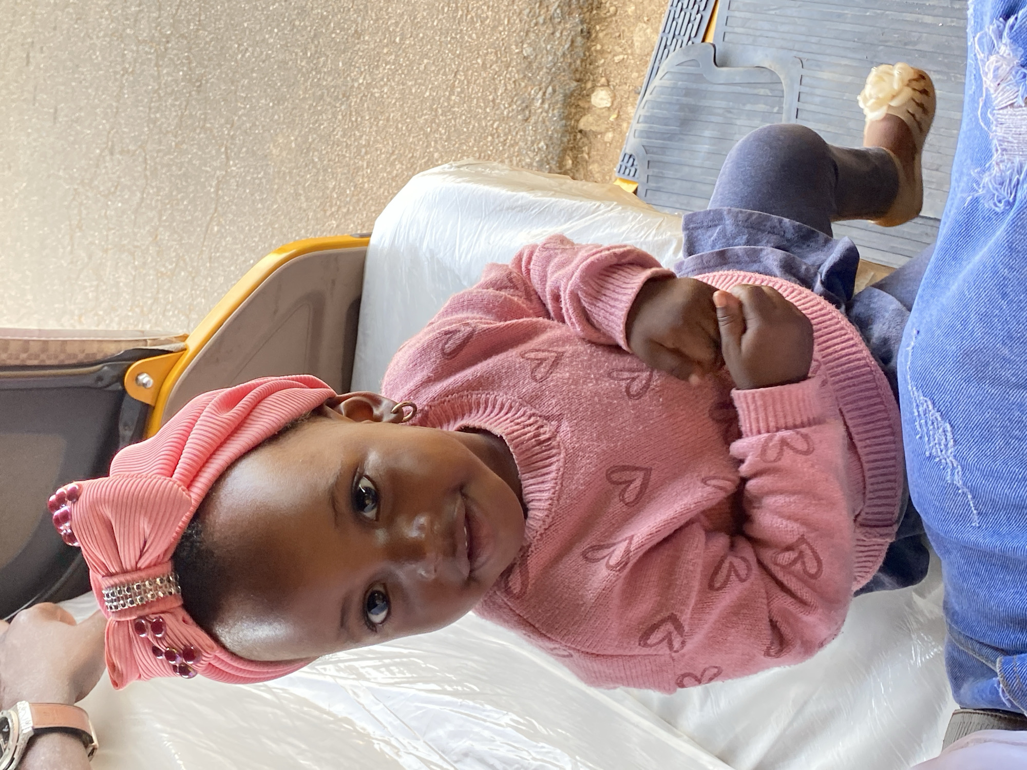 A baby wearing a pink sweater with hearts and a pink head wrap with a jeweled bow is lying on a white plastic-covered surface. Part of a yellow vehicle is visible. An adult wearing jeans and a watch is partially visible in the shot. The location is the city of Vom, Nigeria.
