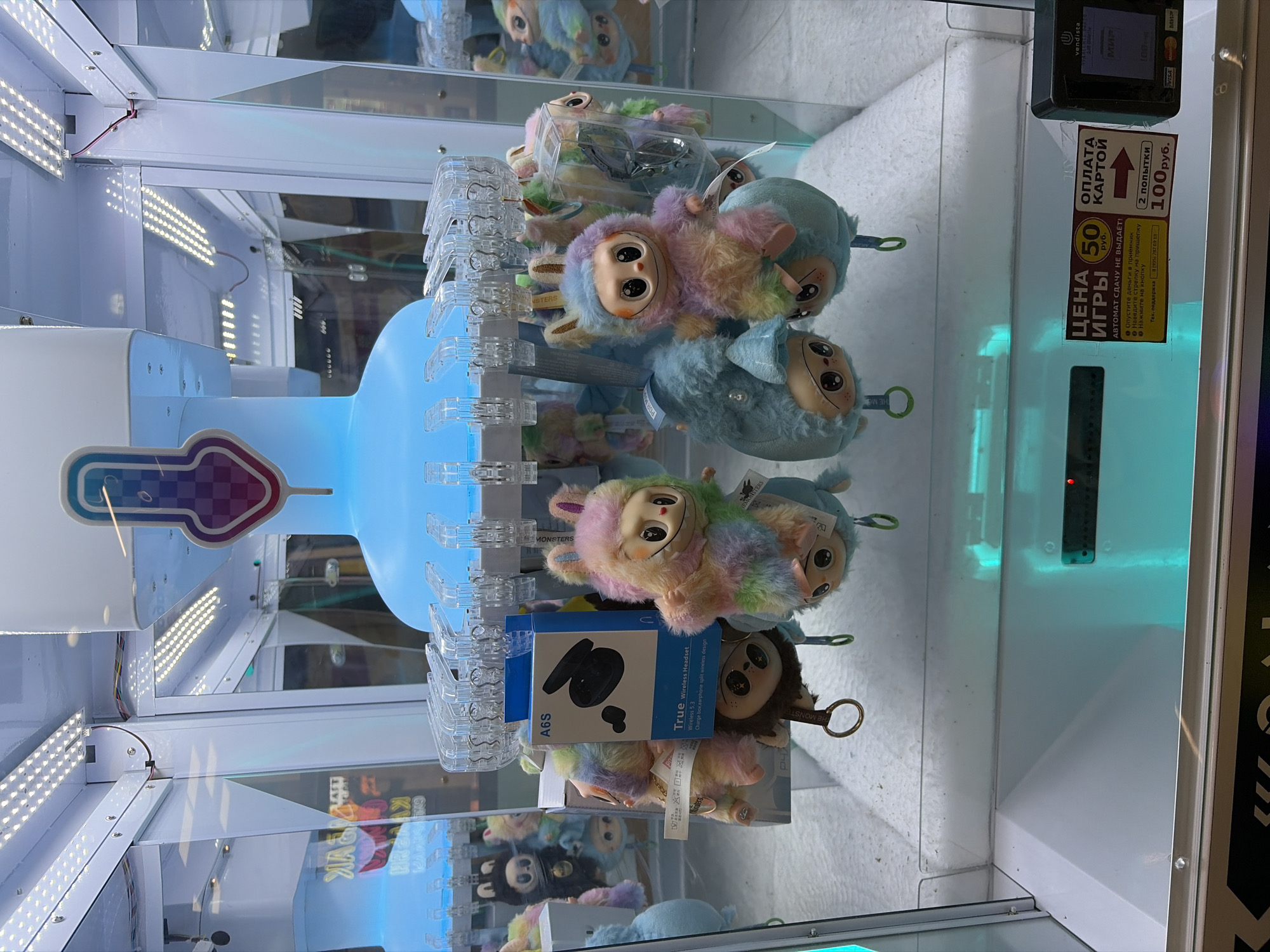 A brightly lit claw machine, likely in Noginsk, Russia, displays an assortment of plush toys. The toys are colorful and have large eyes, some with rainbow-striped fur and rabbit ears. A blue box labeled "A6S True Wireless Headset" is visible among the toys. The machine has clear glass panels, and the interior is illuminated with white LED lights at the top and a teal light at the bottom. A small digital display on a black payment terminal is visible in the lower right corner, showing a transaction amount. A sticker with Russian text, indicating game prices and payment options, is affixed to the machine's lower section. The floor below the machine is white and appears to be a soft, possibly plush, surface.