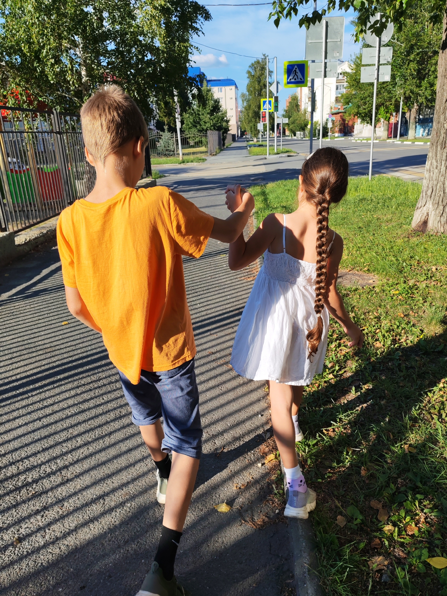 Two children, a boy and a girl, walk hand-in-hand along a sidewalk in an urban environment. The boy, on the left, wears an orange t-shirt and blue striped shorts. The girl, on the right, wears a white sundress and has a long braid. They are walking away from the camera. The sidewalk is paved and is bordered by grass and a tree on the right. In the background, there is a street with crosswalks and traffic signs, as well as buildings and trees. The sun casts long shadows across the sidewalk.