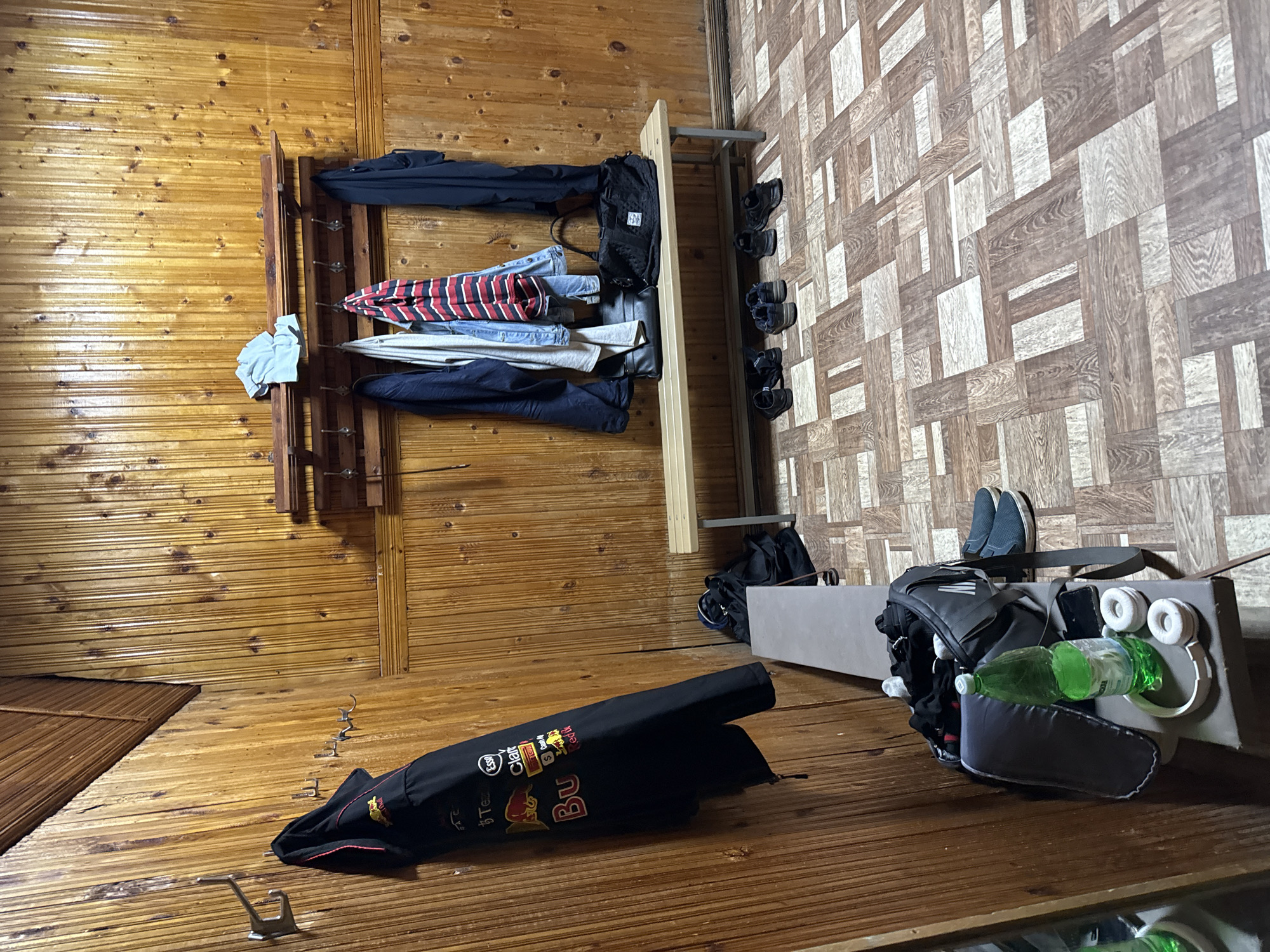 The image depicts a locker room with wood-paneled walls and floor. A coat rack above a wooden bench holds several items of clothing, including a black jacket with Red Bull branding, a blue denim jacket, a striped shirt, and a dark blue jacket. Several bags and pairs of shoes are placed on the floor and the bench. On a lower bench, a green bottle of water, a smartphone, and headphones are visible next to a gym bag. The flooring is a patterned laminate with shades of brown and beige, resembling wood. This scene is located in the city of Kazan, Russia.