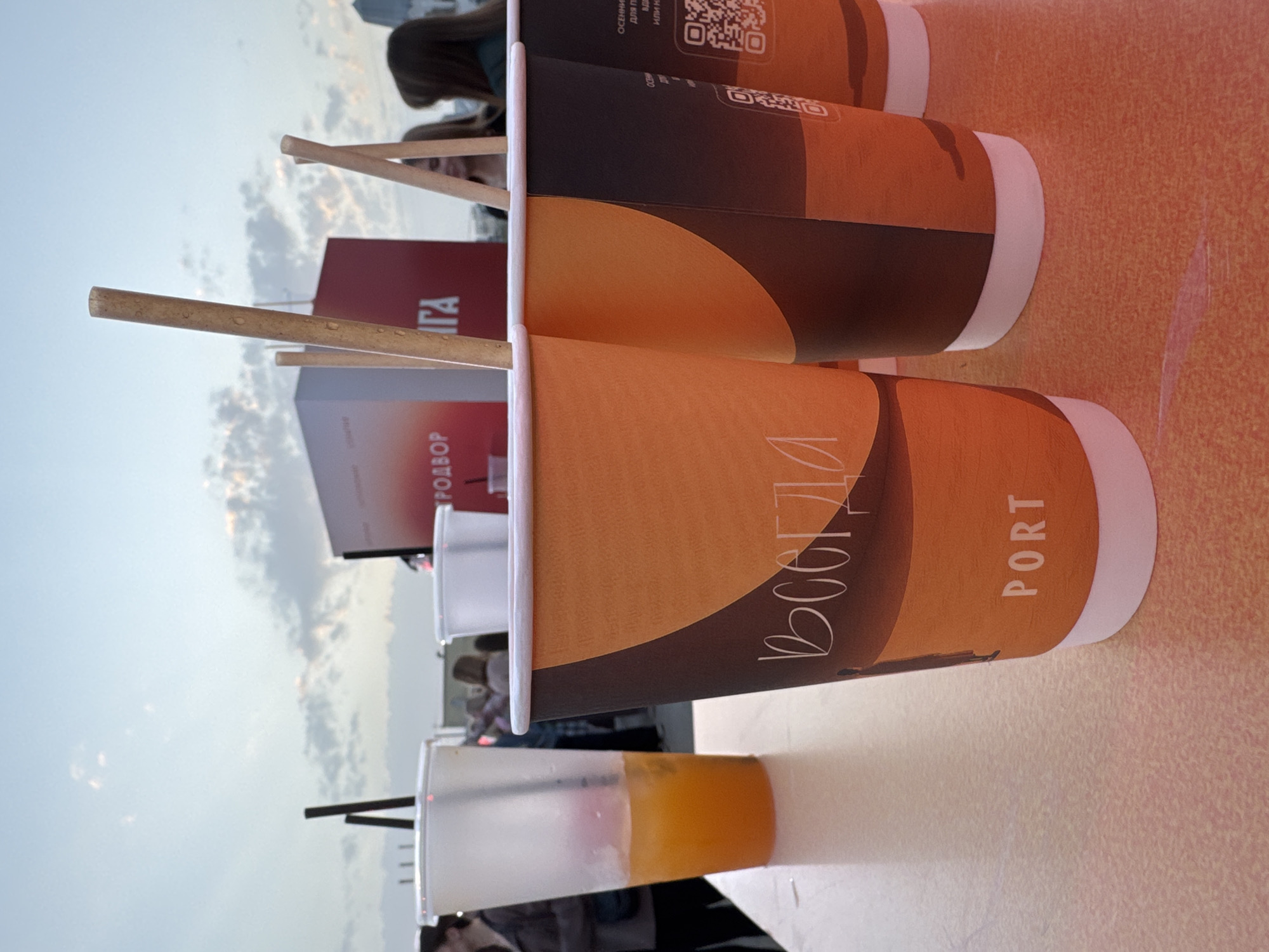 The image depicts a gathering, potentially an outdoor café or event, with focus on a table where a row of beverage cups are arranged.
**Subjects and Objects:**
* **Cups:** There are multiple disposable cups, some containing liquid (potentially juice or a cocktail) and others appear to be coffee cups. The coffee cups have a brown and orange color scheme, with "PORT" and some Russian lettering on them.
* **Stir Sticks/Straws:** Wooden sticks are seen in some of the cups, likely for stirring or sipping beverages.
* **Background:** In the background, there's a hint of a crowd of people. There are also signs or boards with text on them, including the word "РОДВОР" and "ГА" in Cyrillic script.
**Environment:**
* The location appears to be outdoors, given the sky visible in the background. It seems to be taken during the daytime.
* Based on the Cyrillic text and information on the cups, it is likely taken in a location where Russian is spoken.
**City:**
* **Saratov, Russia:** I am unable to determine if the photograph was taken in Saratov specifically.