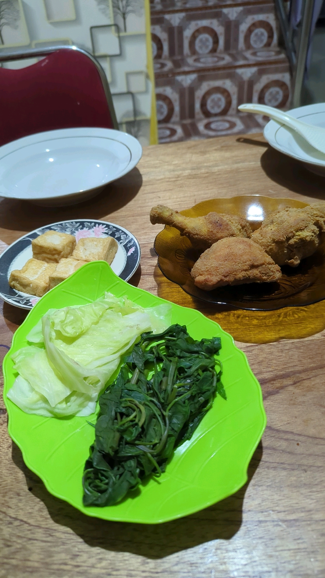 A close-up, eye-level shot captures a meal laid out on a wooden table. In the foreground, a bright green leaf-shaped plate holds cooked leafy green vegetables and blanched cabbage. To the left, a smaller plate with a floral rim displays four pieces of fried tofu. To the right, a brown glass plate holds three pieces of fried chicken, including a drumstick and two other pieces. In the background, a white bowl with a golden rim sits on the table, and beyond that, a set of stairs with patterned tiles is visible. The overall scene suggests a home-cooked meal served in Baroqah, Indonesia.
