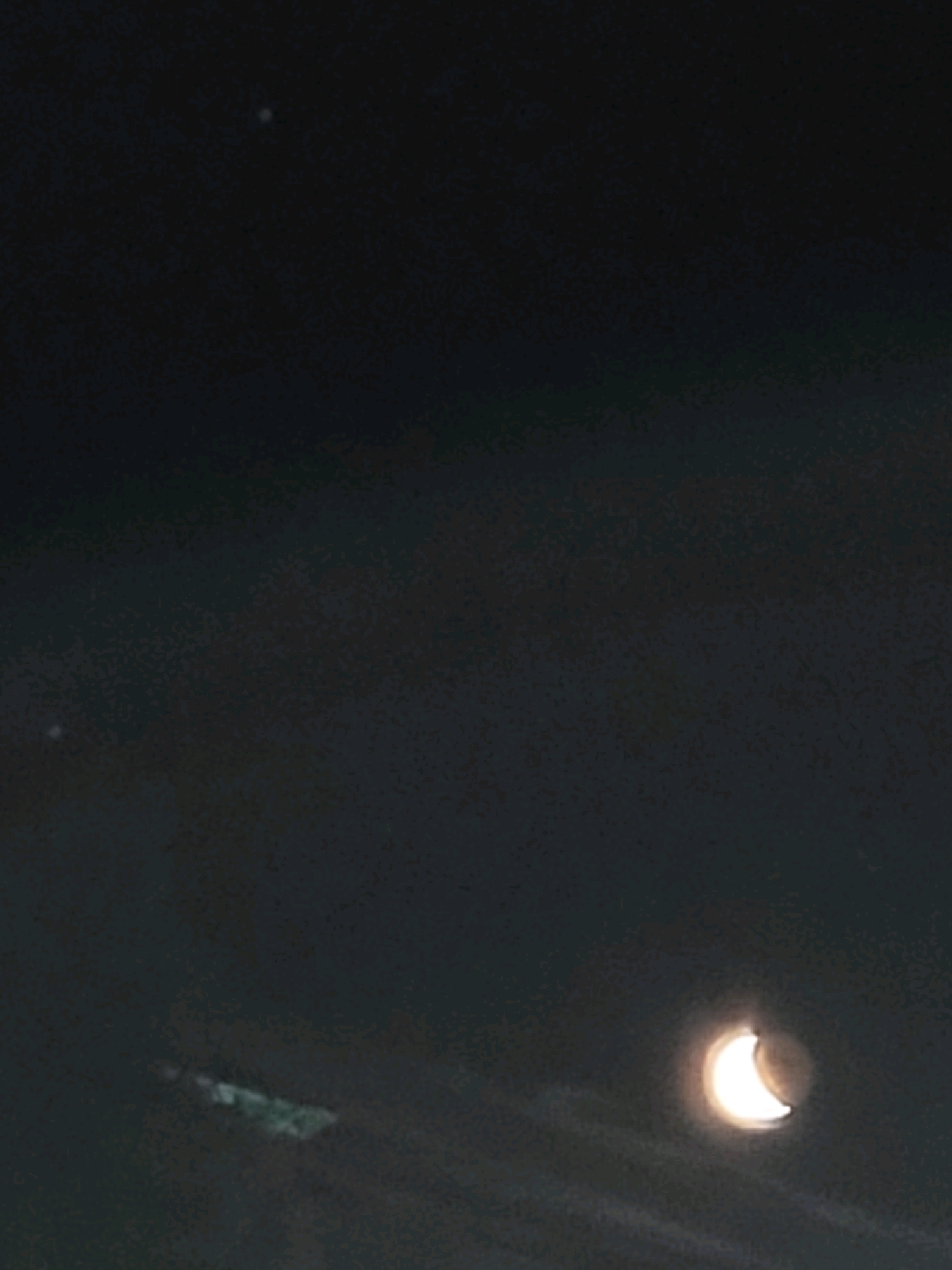 The image captures a nighttime sky with a crescent moon prominently featured in the lower right corner, emitting a bright glow. There are at least two faint stars visible in the upper portion of the frame. A blurry, indistinct shape, possibly a structure or cloud formation, is visible in the lower left quadrant. The overall environment appears to be outdoors, likely in a semi-rural or natural setting, given the lack of urban development in the visible areas and the presence of celestial bodies. There are no people or specific objects identifiable that would pinpoint the location within Ozolnieki, Latvia, beyond the general assumption of an outdoor setting at night.