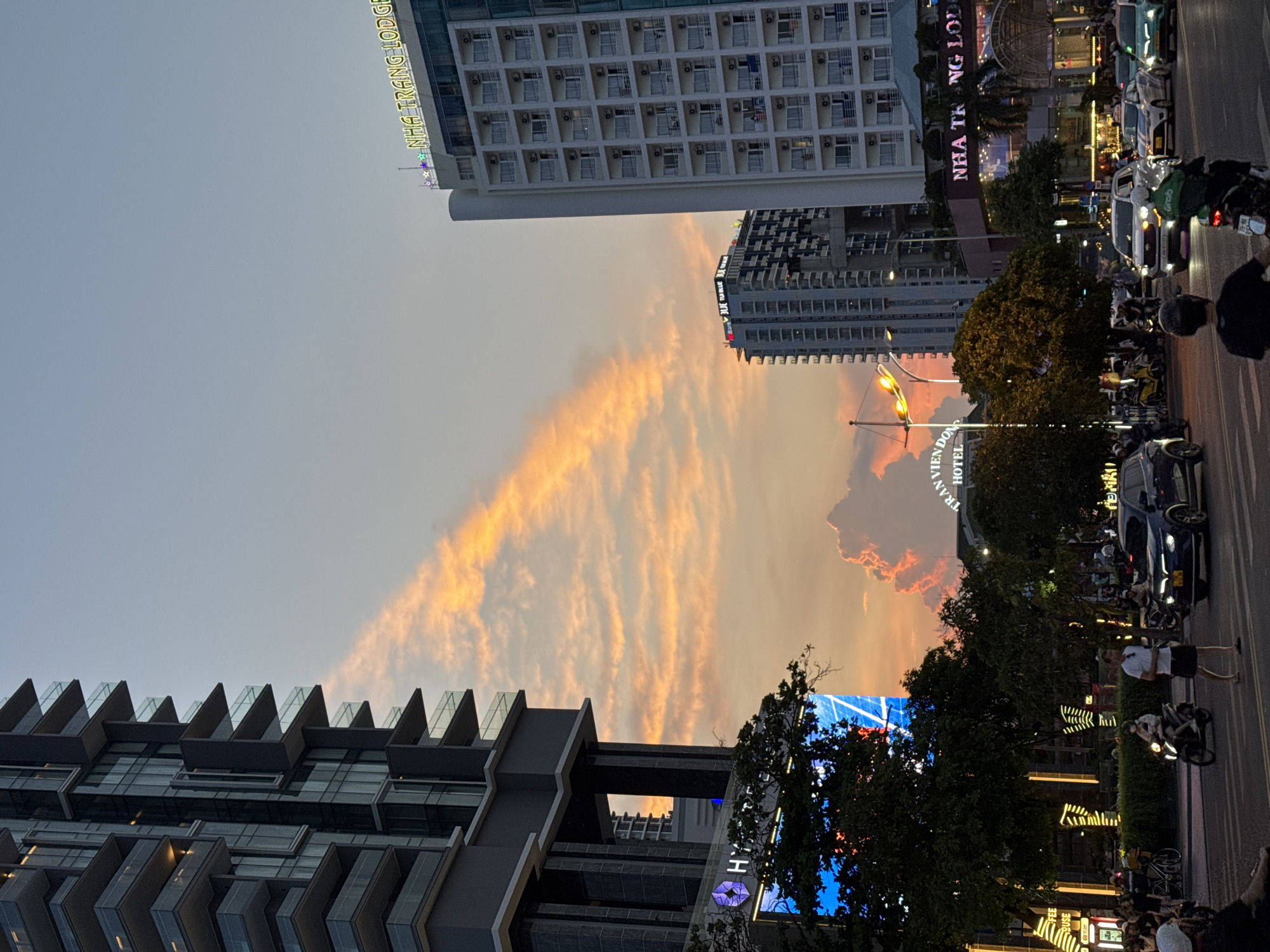 This image captures a street scene in Nha Trang, Vietnam, during a twilight hour. Tall modern buildings dominate the skyline, one on the left with dark gray siding and numerous balconies, and another on the right, a white multi-story structure with rows of windows, identified by a neon sign as "NHA TRANG LODGE." The sky is painted with dramatic sunset colors, featuring streaks of orange and pink clouds, with a particularly vibrant, bulbous cloud formation visible in the distance.
The street below is bustling with activity. Cars and motorcycles are navigating the road, with a dark blue car in the foreground and a white SUV with "Grab" branding further back. Several people are visible on foot, including a man in a white t-shirt and black shorts crossing the street, and others walking along the sidewalk. Some individuals are riding motorcycles, one of whom is wearing a helmet and a green jacket. Trees line the street, providing a dark contrast to the illuminated buildings and signs. A large digital screen displaying a blue and white graphic is visible on a building to the left, next to a tree. Neon signs with festive lighting also illuminate the storefronts. The overall impression is a vibrant urban environment at dusk, with the natural beauty of the sunset juxtaposed against the modern architecture and street life.
