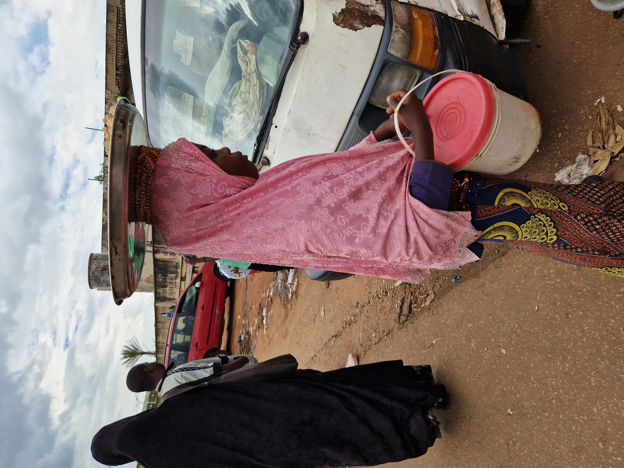 In Jos, Nigeria, a young girl, her head covered in a pink hijab adorned with floral patterns and small rhinestones, carries a tray of food items balanced on her head. She is wearing a vibrant, patterned skirt and holding a white bucket with a red lid in her right hand. Beside her stands a person in a black hijab. In the background, a red car is parked on a dirt road, and a white van with significant rust and damage is partially visible. The scene unfolds under a cloudy sky.