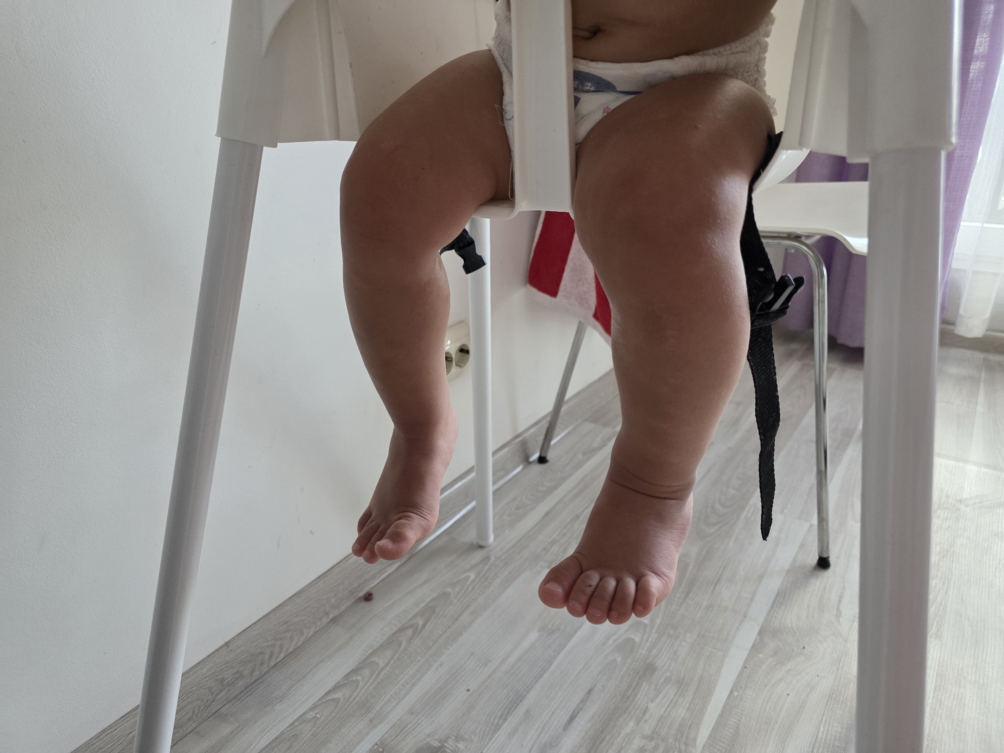 The image shows the lower body of a baby sitting in a white high chair. The baby is wearing a diaper and has chubby legs and feet. The high chair has white plastic legs and a white seat. A black strap is hanging down from the side of the seat. The background includes a light-colored wall and a light wooden floor. To the right, a sheer purple curtain is visible. A power outlet is visible on the wall behind the high chair. The scene appears to be indoors, likely in a home environment. The location of Sochi, Russia is not discernible from the image.