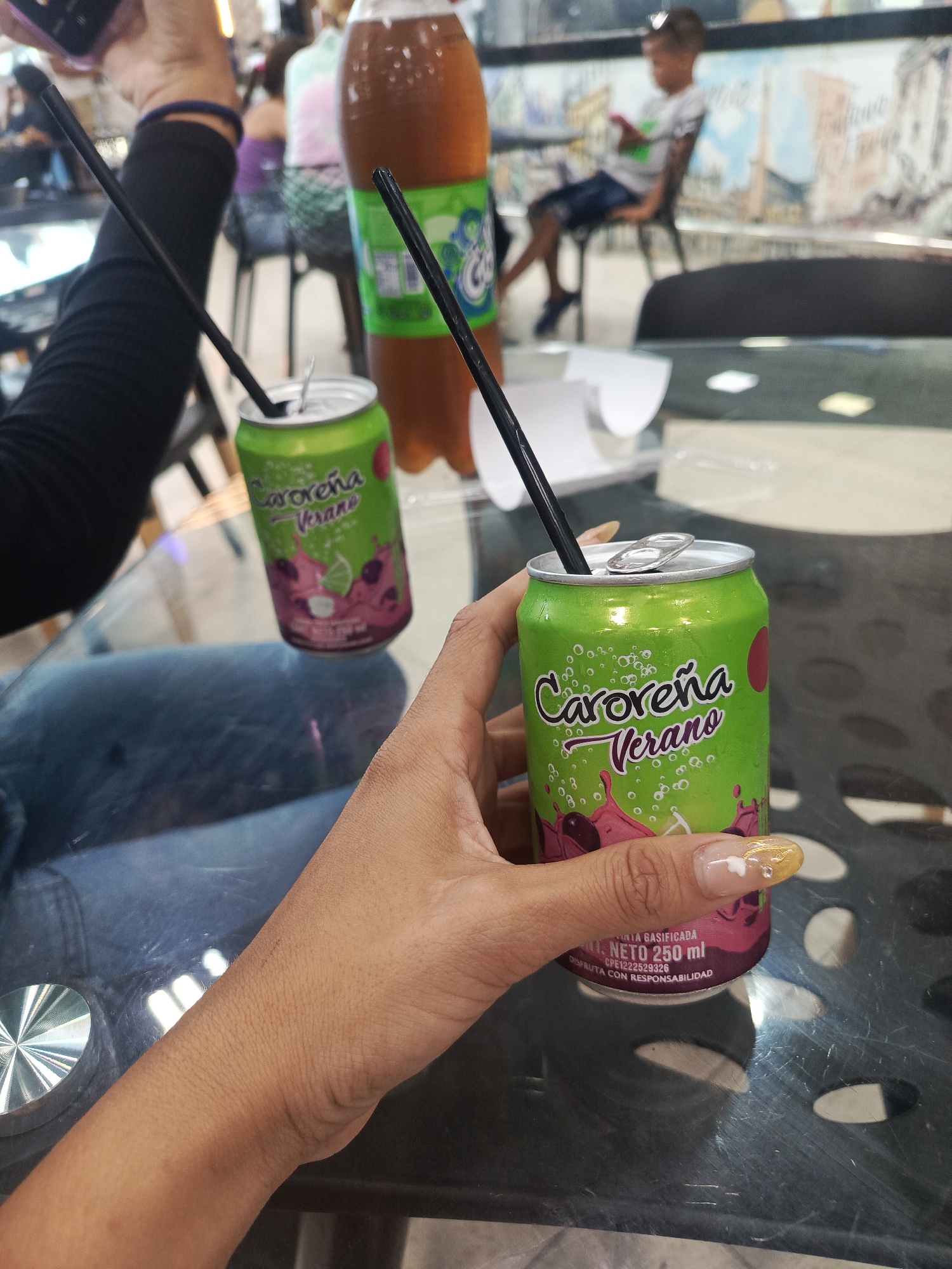 The image depicts a table in what appears to be a public space, possibly a cafeteria or food court. On the table are two cans of "Caroreña Verano" sparkling grape juice, each with a black straw inserted. There is also a bottle of a similar-colored drink and a white container nearby. A person's hand is holding one of the cans. Another person's arm is visible to the left, wearing a black sleeve. In the background, there are other people seated and standing. Based on the branding of the drinks, specifically "Caroreña," which is associated with Venezuela, and the description "Verano," which translates to "Summer," it is likely the location is in Venezuela. However, the exact location, specifically within the city of Juan Griego, cannot be definitively determined from the visual information provided in the image.