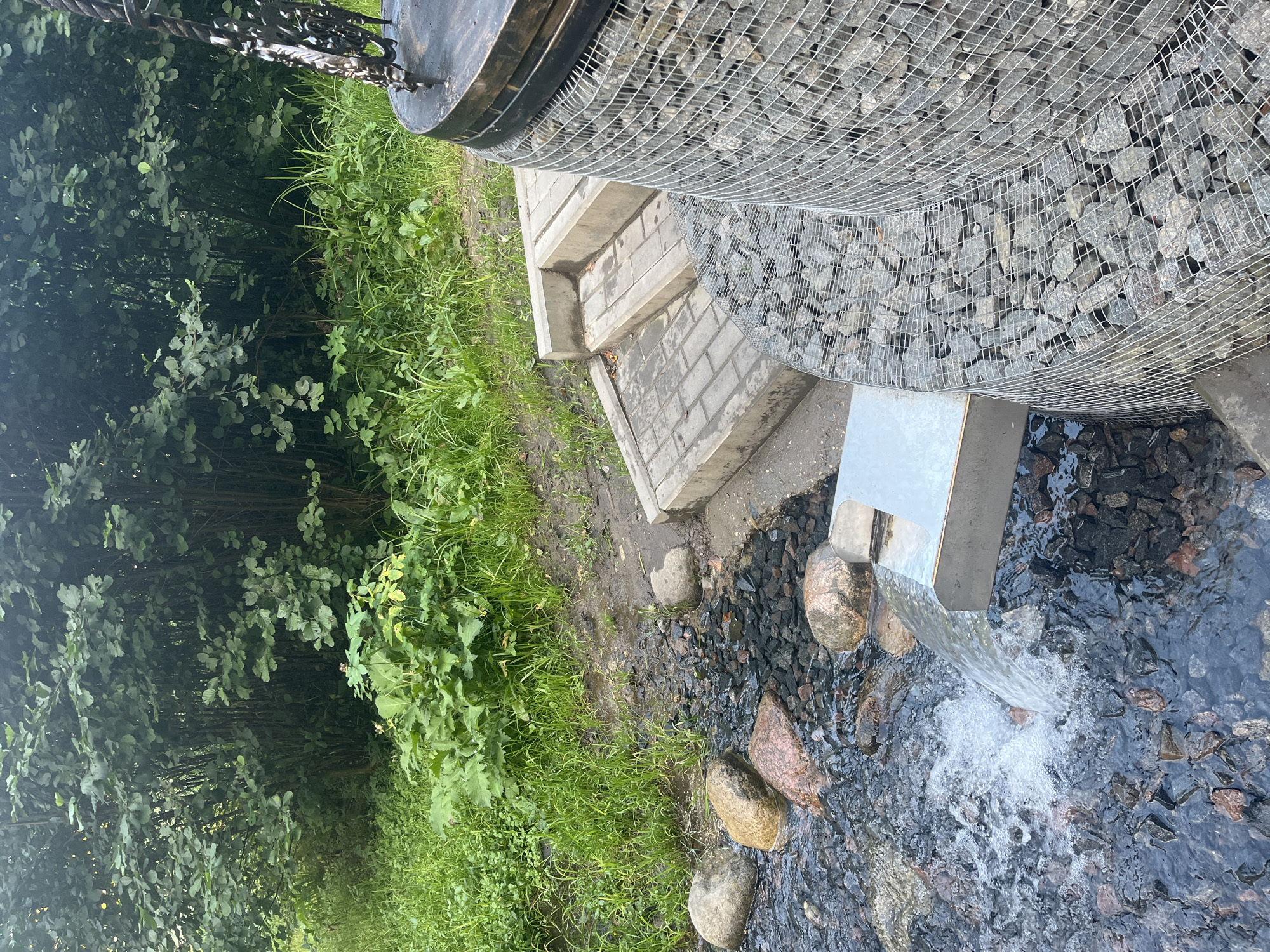 In the city of Lahojsk, Belarus, a small stream flows over dark gravel and large, smooth rocks. The water cascades from a shiny metal spout into the stream, creating a foamy splash. To the right of the stream, a rounded structure, partially enclosed by a wire mesh filled with gray stones, rises upwards. Adjacent to this structure, a set of concrete steps leads up from a paved walkway. Behind the stream and steps, lush green grass and dense foliage from trees create a natural backdrop. The scene appears to be an outdoor, landscaped area.