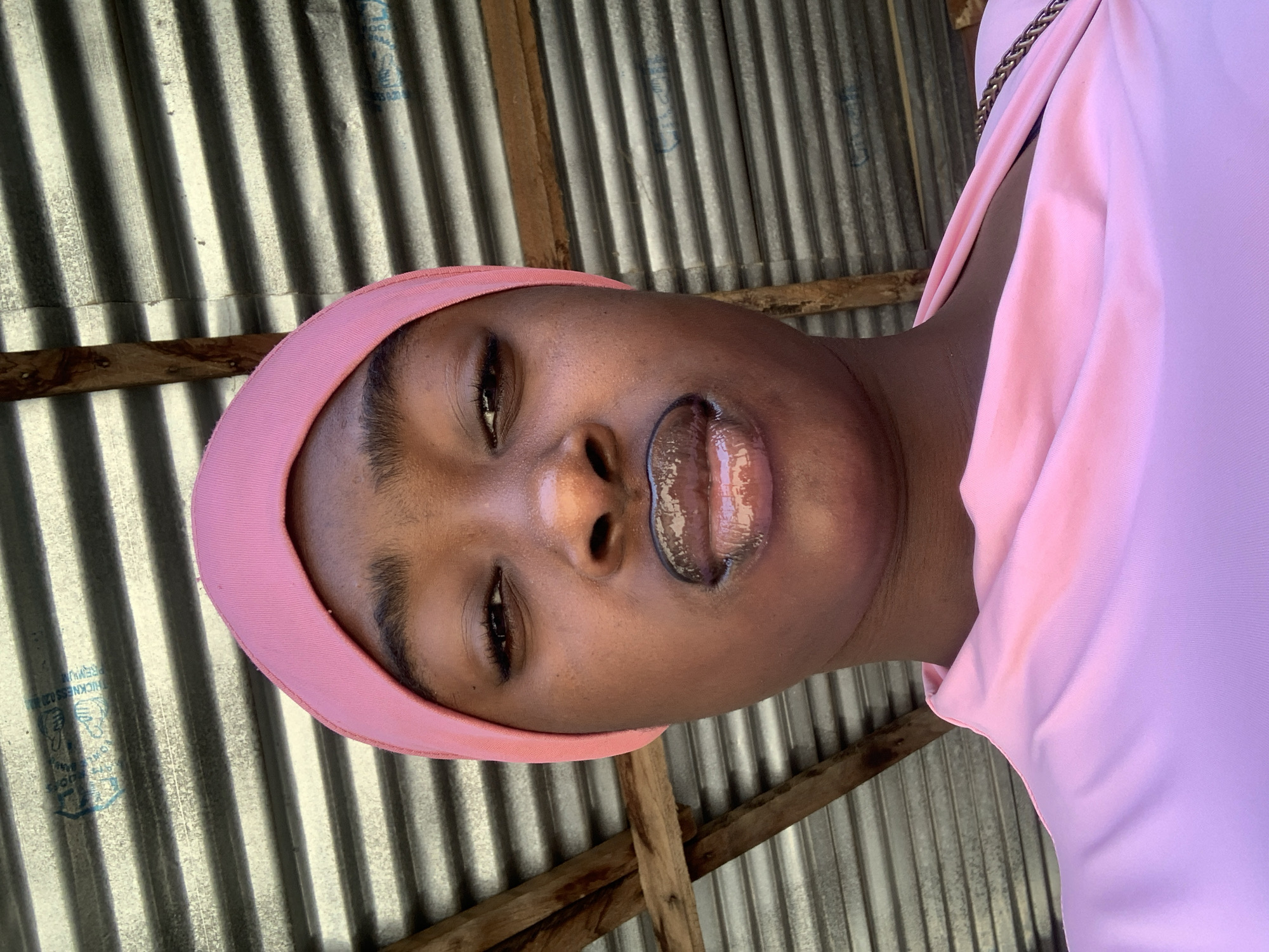 A close-up, tilted portrait shows a woman wearing a pink head wrap and a pink top with a gold chain around the neck. She has dark brown eyes and shiny lips. The background is corrugated metal and wooden beams. The location is possibly Rafin Par, Nigeria.