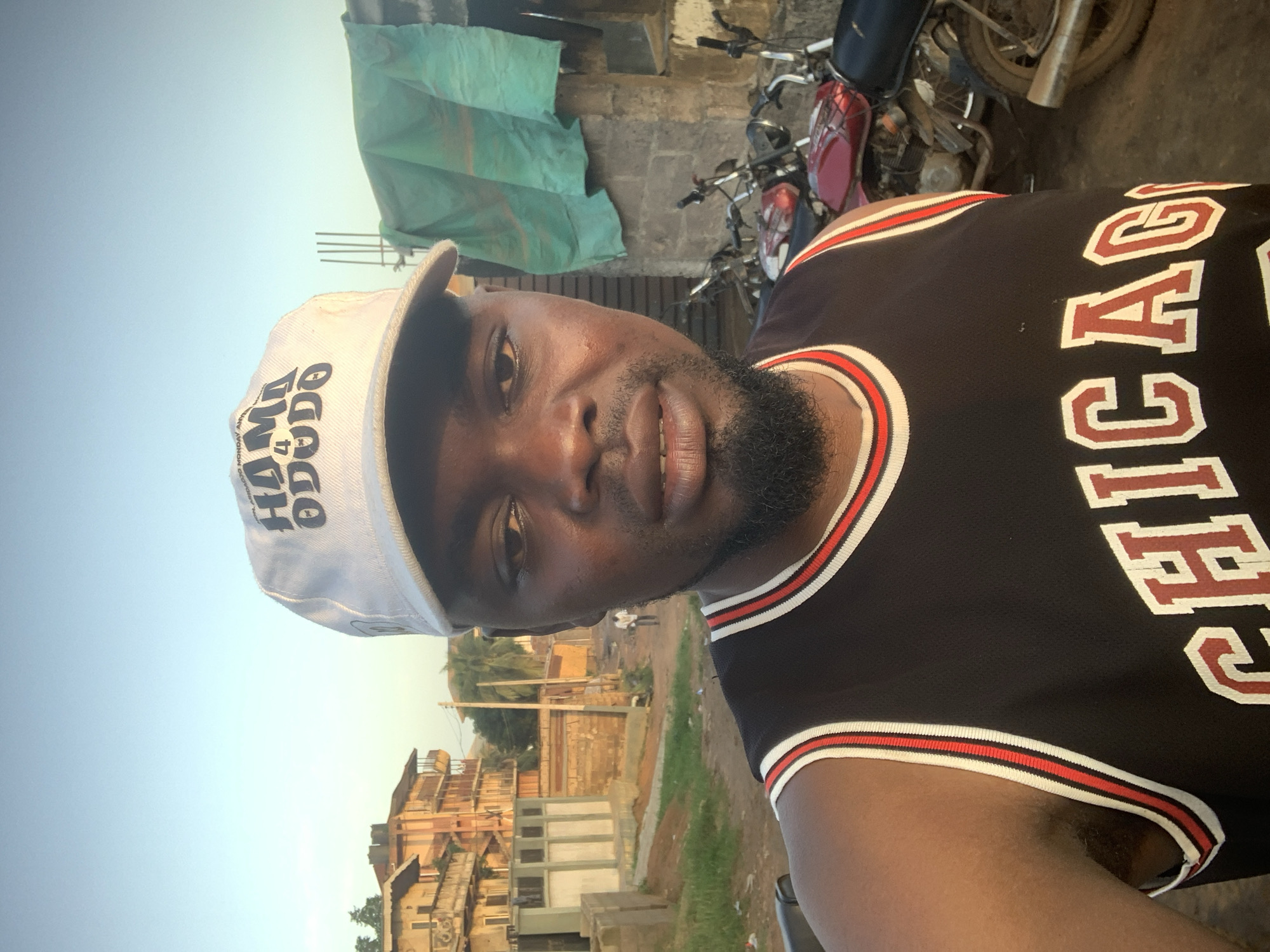 A dark-skinned man wearing a white baseball cap with black lettering and a black sleeveless basketball jersey with red and white trim is taking a selfie outdoors. The man has a beard and is looking directly at the camera. The background shows a street scene in Shagamu, Nigeria, with several buildings, including a multi-story residential building with balconies and a smaller brick structure covered with a teal tarp. Several motorcycles are parked in front of the brick structure. The sky is clear and blue, suggesting it is daytime.
