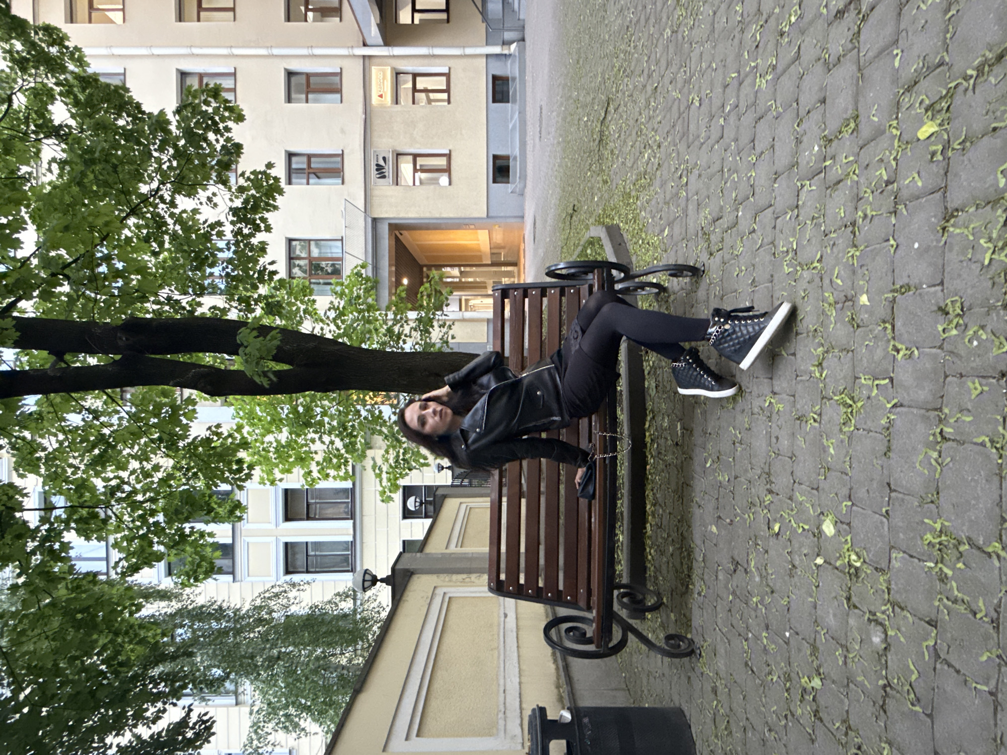 The image depicts a woman reclining on a park bench in Saint Petersburg, Russia. She is wearing a black leather jacket, black leggings, and black sneakers with white soles. The bench is made of wood and cast iron. The background includes a building with multiple windows, a tree with green leaves, and a cobblestone walkway covered with fallen leaves. A trash can is located at the bottom of the frame.